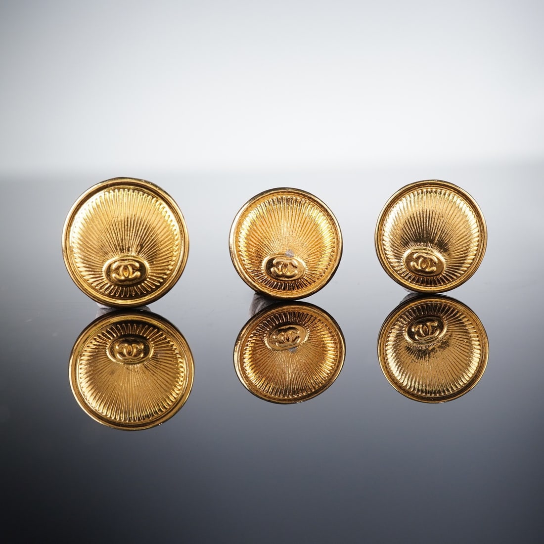 A Set of Three Chanel Vintage Gold-Toned CC Radiated Buttons: A set of three Chanel vintage gold-toned buttons, featuring the iconic interlocking double 'C' logo at the center, surrounded by radiating etched lines creating a sunburst pattern. The reverse of each