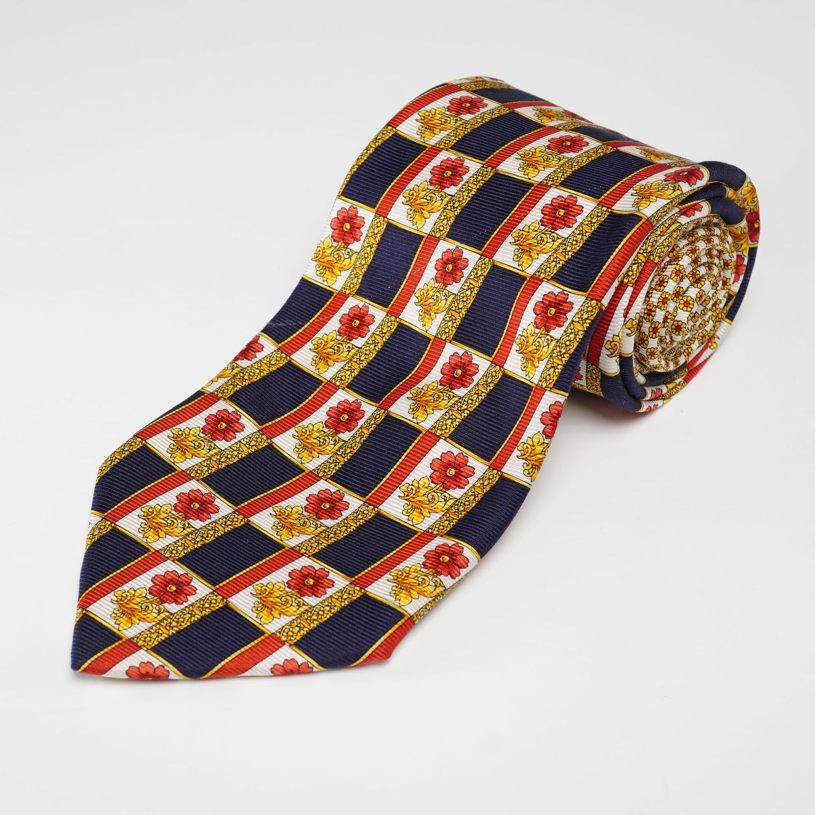 GIANNI VERSACE 100% Silk Necktie with Grid and Floral Pattern: A GIANNI VERSACE 100% silk necktie. The tie features a repeating grid pattern with dark blue square cells outlined in red and gold, alternating with white squares containing elaborate gold floral or b