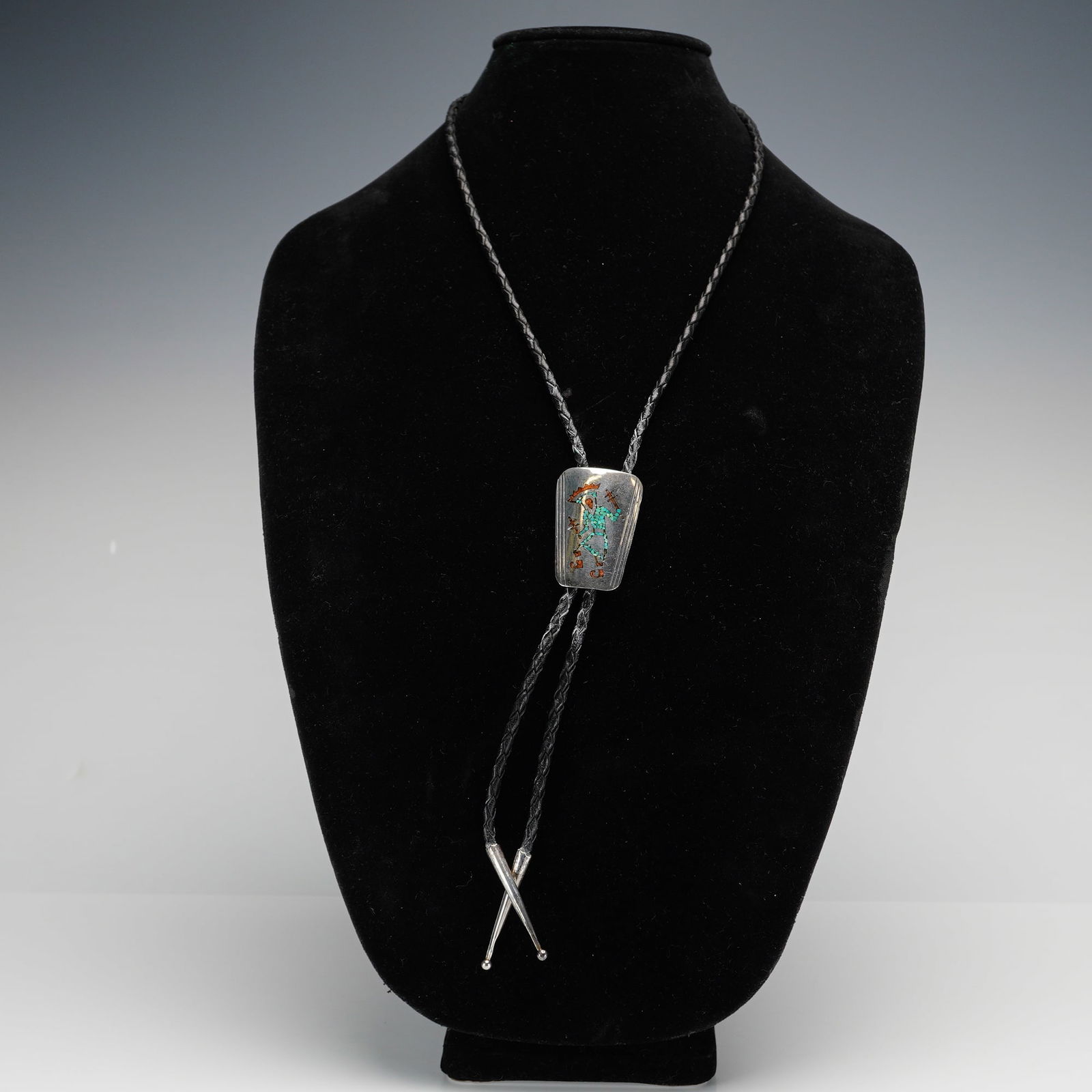 Bennett Navajo Sterling Silver Turquoise and Coral Bolo Tie: This Navajo sterling silver bolo tie features a rectangular slide with rounded corners, decorated with a central dancer figure crafted from turquoise and coral chip inlay. The figure is flanked by ver