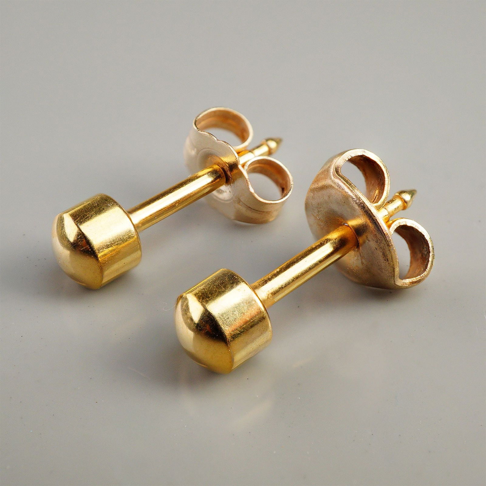 A Pair of 14K Yellow Gold Minimalist Stud Earrings: A pair of minimalist stud earrings constructed of 14K yellow gold. Each earring features a capped cylindrical stud head attached to a straight post, secured by a butterfly or friction back. The design