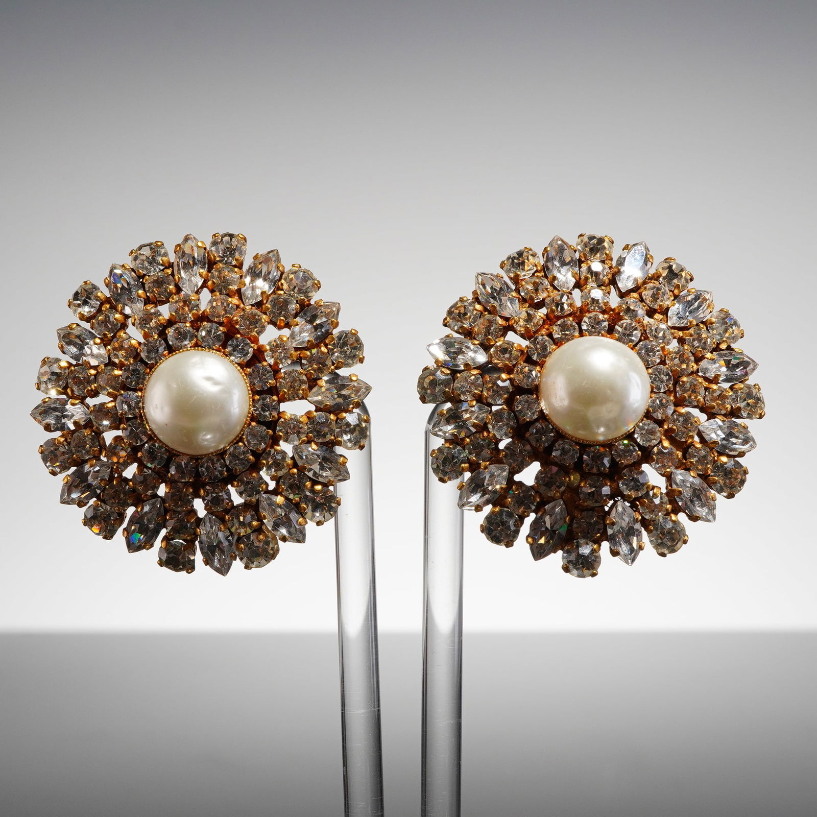 Pair of Chanel Faux Pearl Rhinestone Camellia Earrings: Each earring is executed in a gold-tone base metal setting designed in an elaborate sunburst or camellia floral motif. The center of each piece features a large circular faux pearl cabochon, surrounde