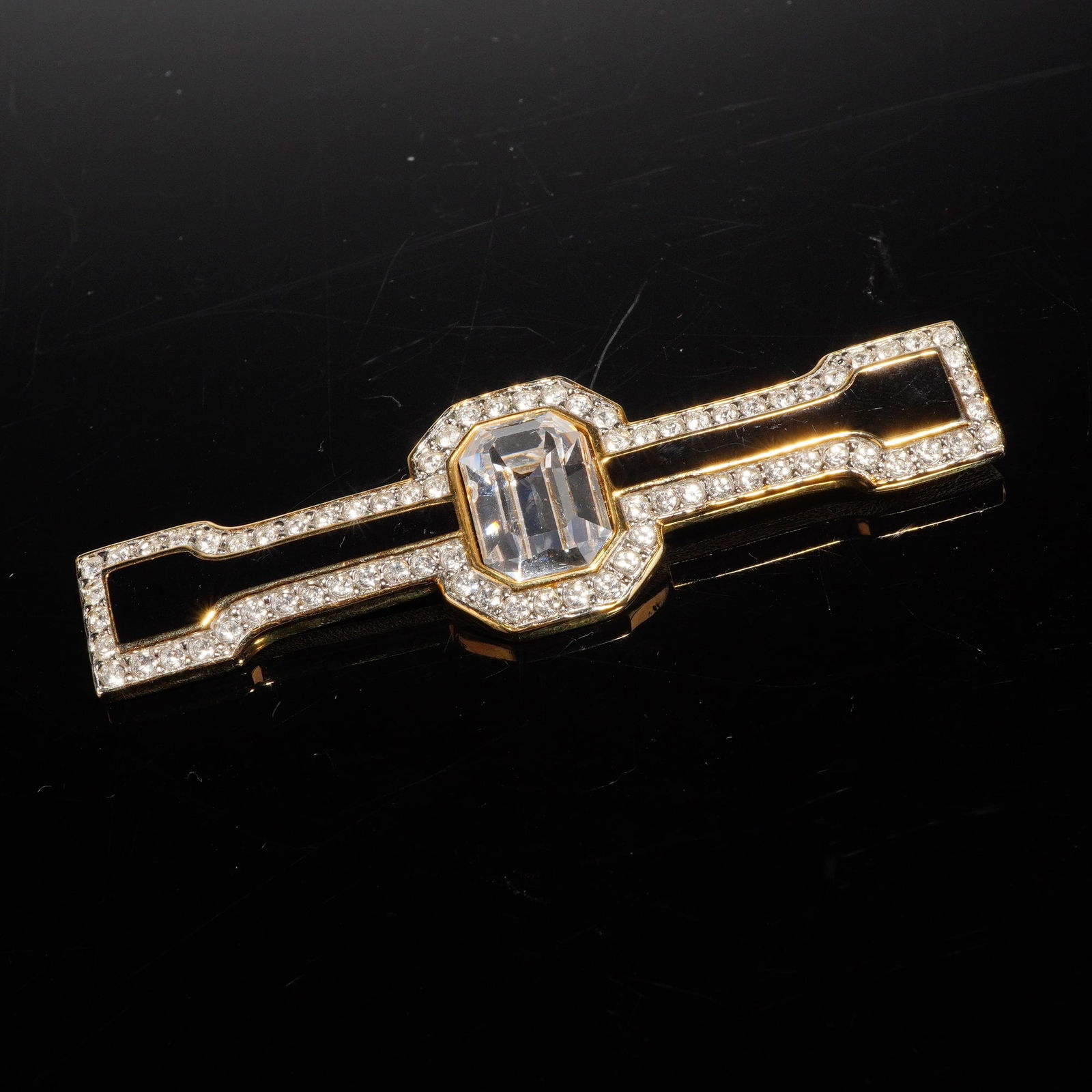 Vintage Art Deco Style Gold Toned Rhinestone and Enamel Brooch: A single bar brooch constructed from gold toned metal, featuring an Art Deco-inspired geometric design. The piece is characterized by a central, rectangular, step-cut colorless simulant stone, surroun