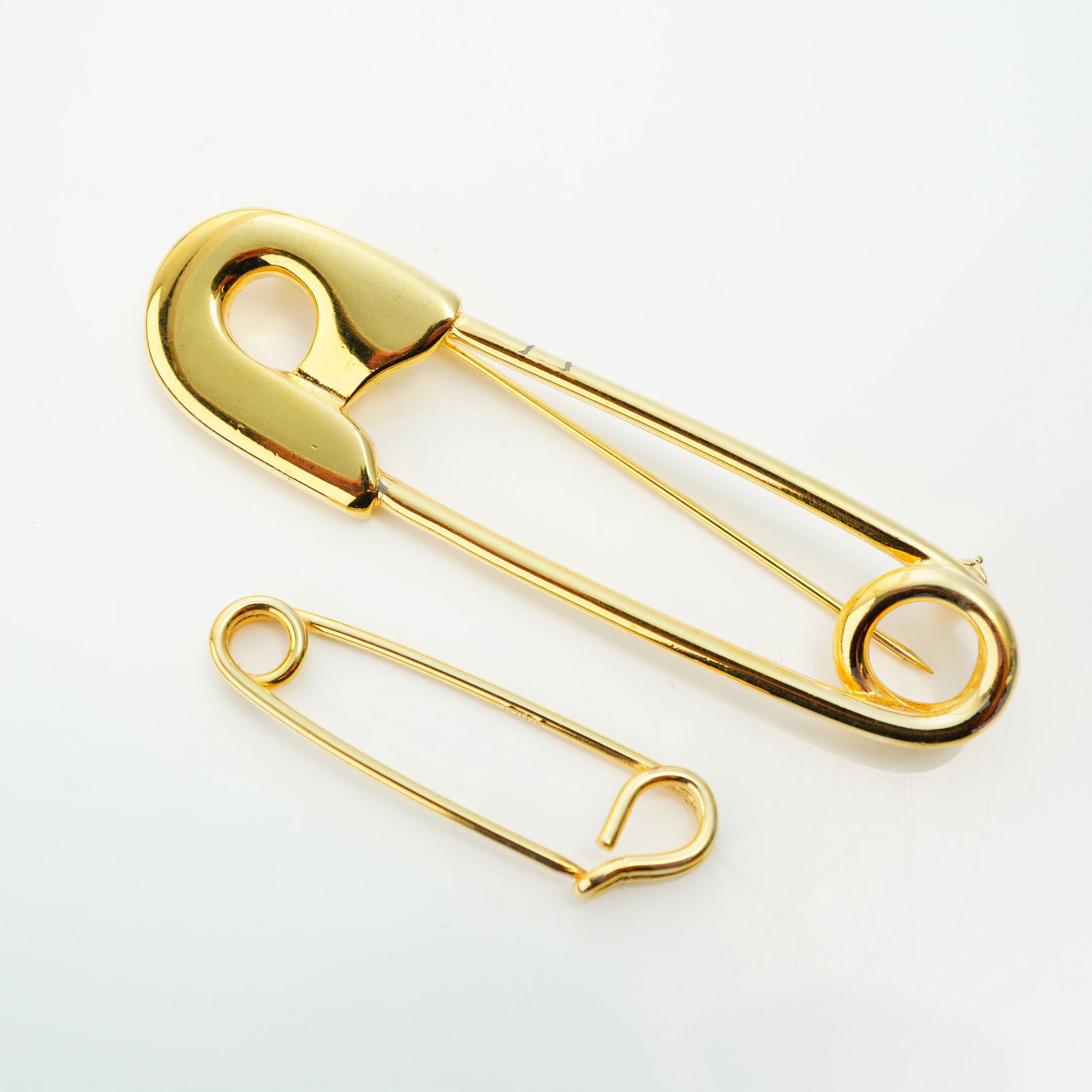 2pc Gold Tone Custom Jewelry Safety Pin Brooches: A duo of custom jewelry gold tone safety pin brooches of differing sizes, both finished in a polished yellow metal. The larger item functions as a costume jewelry brooch, featuring an oversized safety
