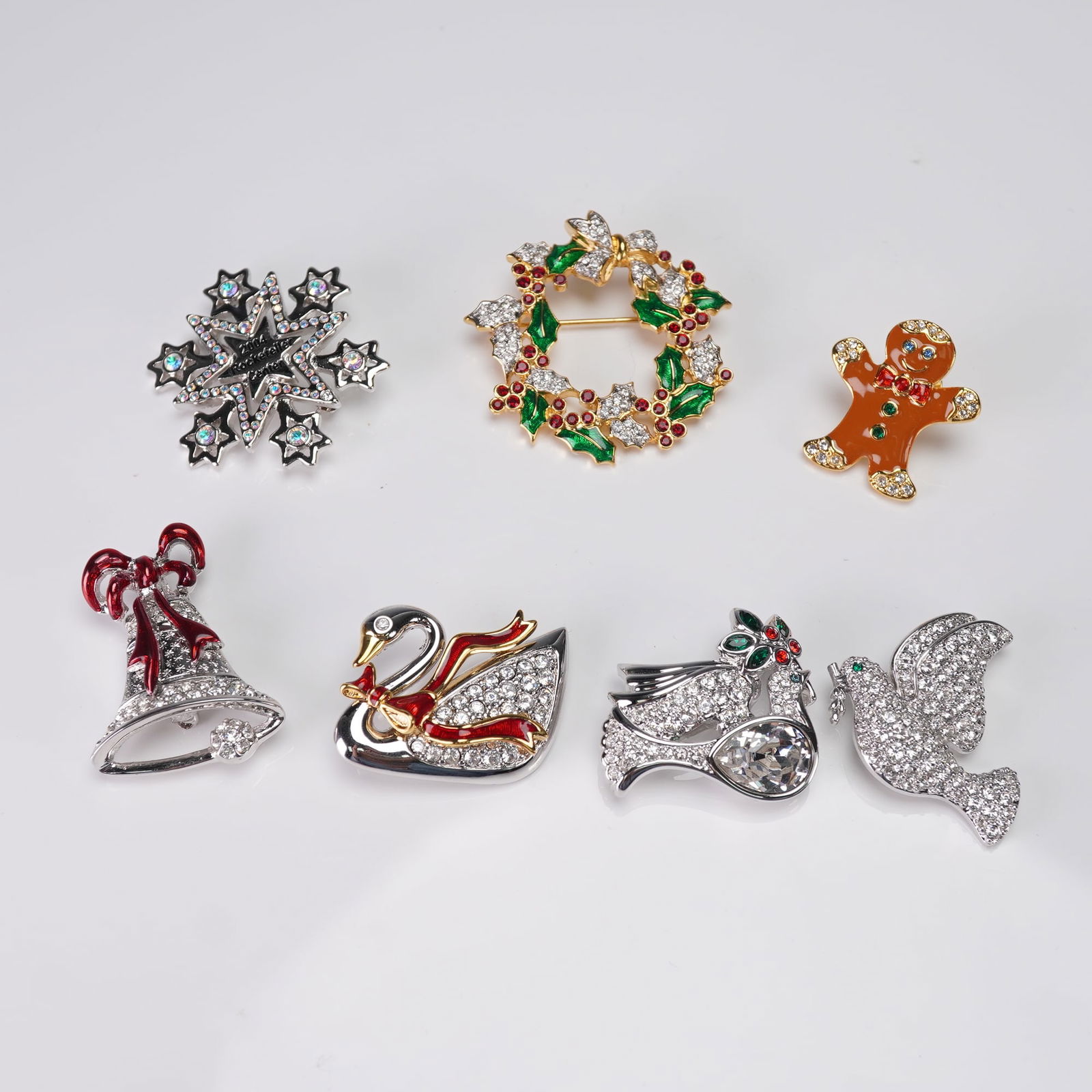 7pc Collection of Swarovski Holiday-Themed Costume Brooches: A collection of seven Swarovski holiday-themed costume brooches. The group includes a swan brooch with a red ribbon, and a Christmas wreath brooch, the remaining five brooches feature designs such as