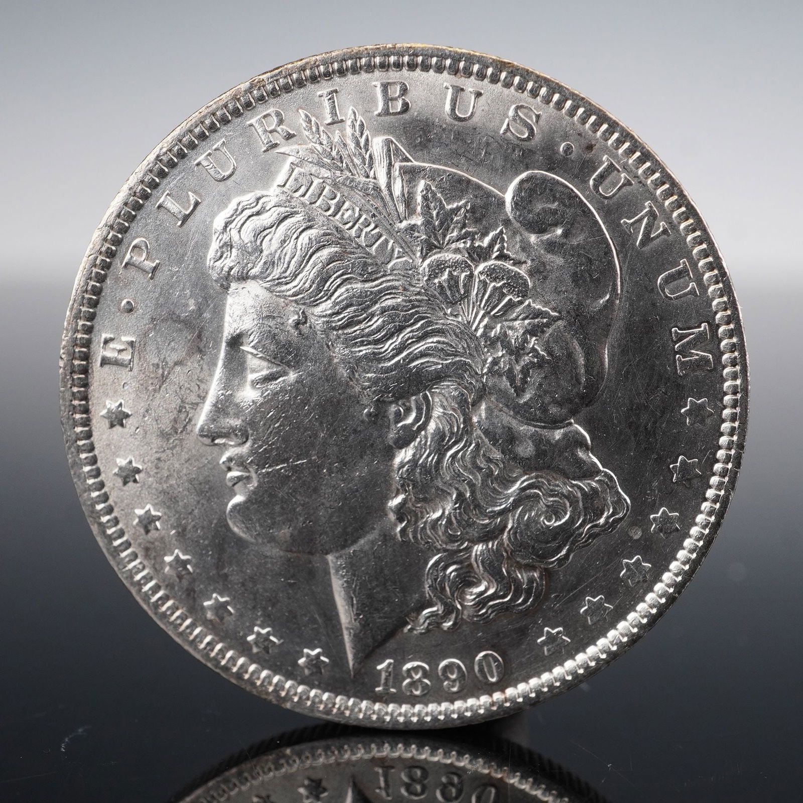 1890-O Morgan Silver Dollar Coin: Struck at the New Orleans Mint and is composed of .900 silver and .100 copper. The obverse features a detailed left-facing portrait of Liberty wearing a Phrygian cap adorned with wheat and cotton, sur