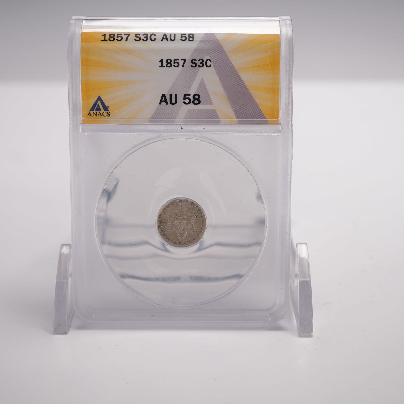 1857 Trime 3-Cents ANACS Graded AU 58: The obverse displays the American shield set within a six-pointed star. The reverse features the Roman numeral III centered within a large stylized letter C, surrounded by thirteen stars. Struck in