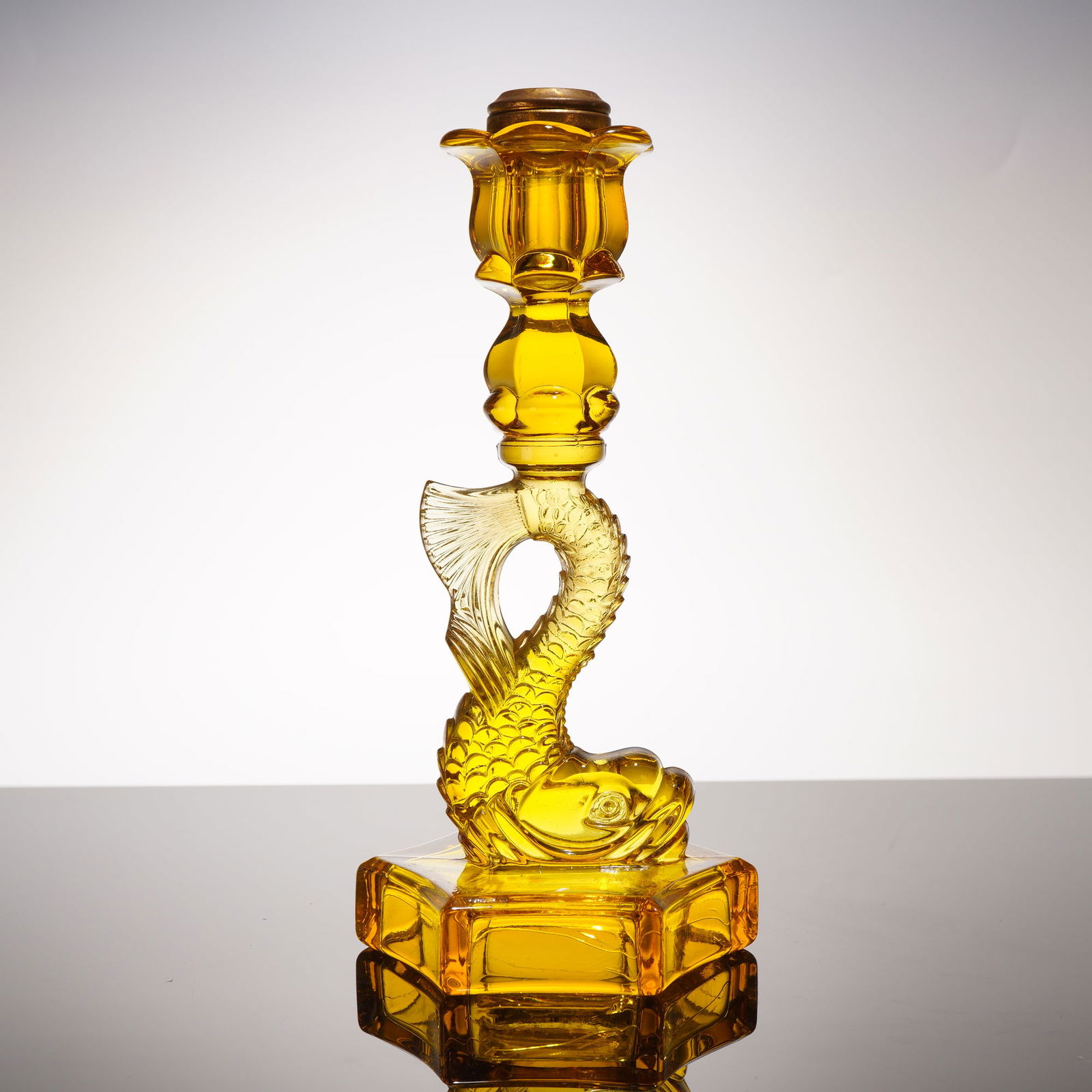 Westmoreland Glass Amber Glass Dolphin Candlestick: This amber pressed glass candlestick features a brass rim encircling the candle hole. The body's design follows the classic Early American Pattern Glass dolphin motif, characterized by a stylized fish