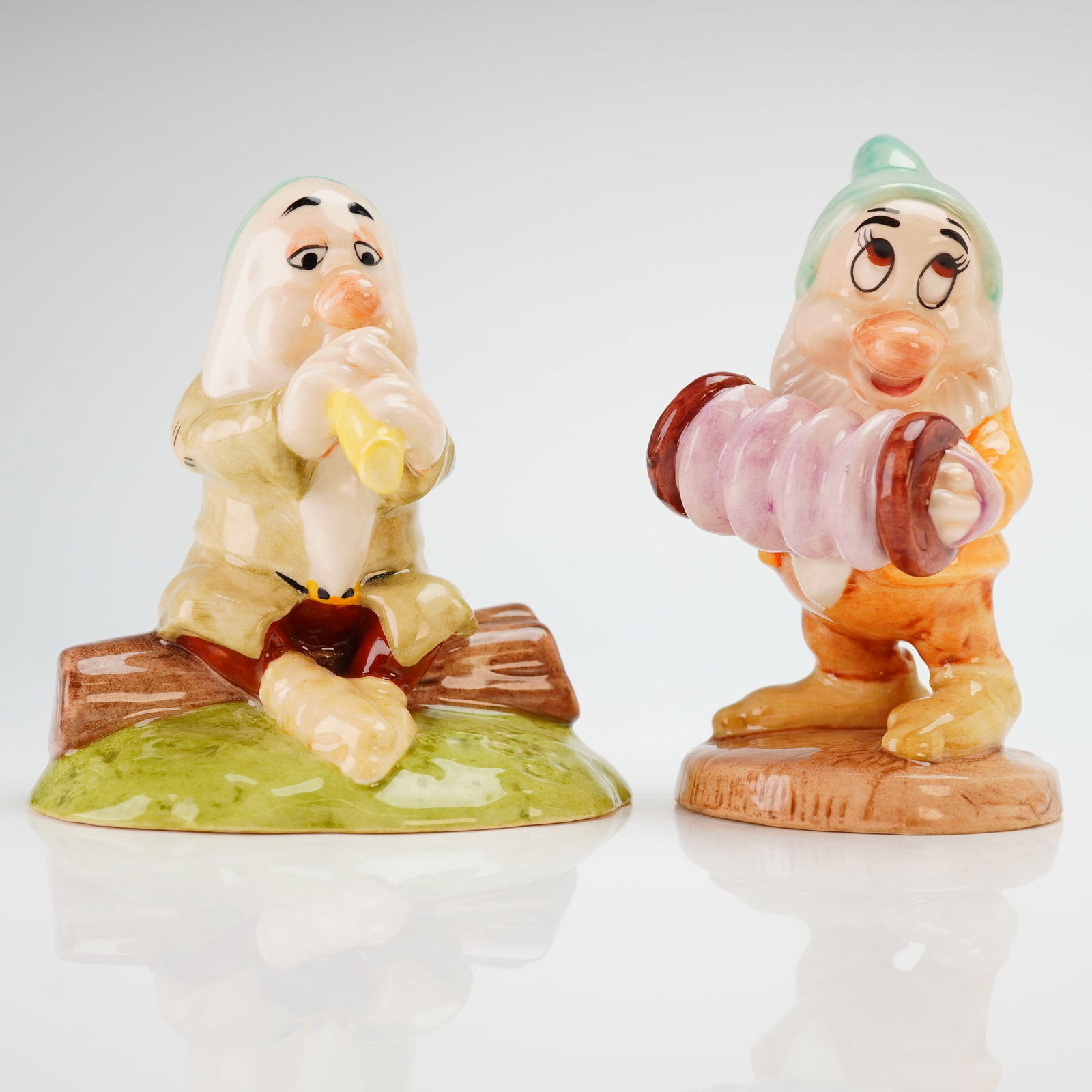 2pc Royal Doulton Disney Snow White Figurines, Bashful's Melody & Sleepy: Each figurine is constructed from glazed porcelain with a high-gloss finish in vibrant multi-colored tones. The figurine titled Bashful's Melody depicts the character playing a concertina, while the
