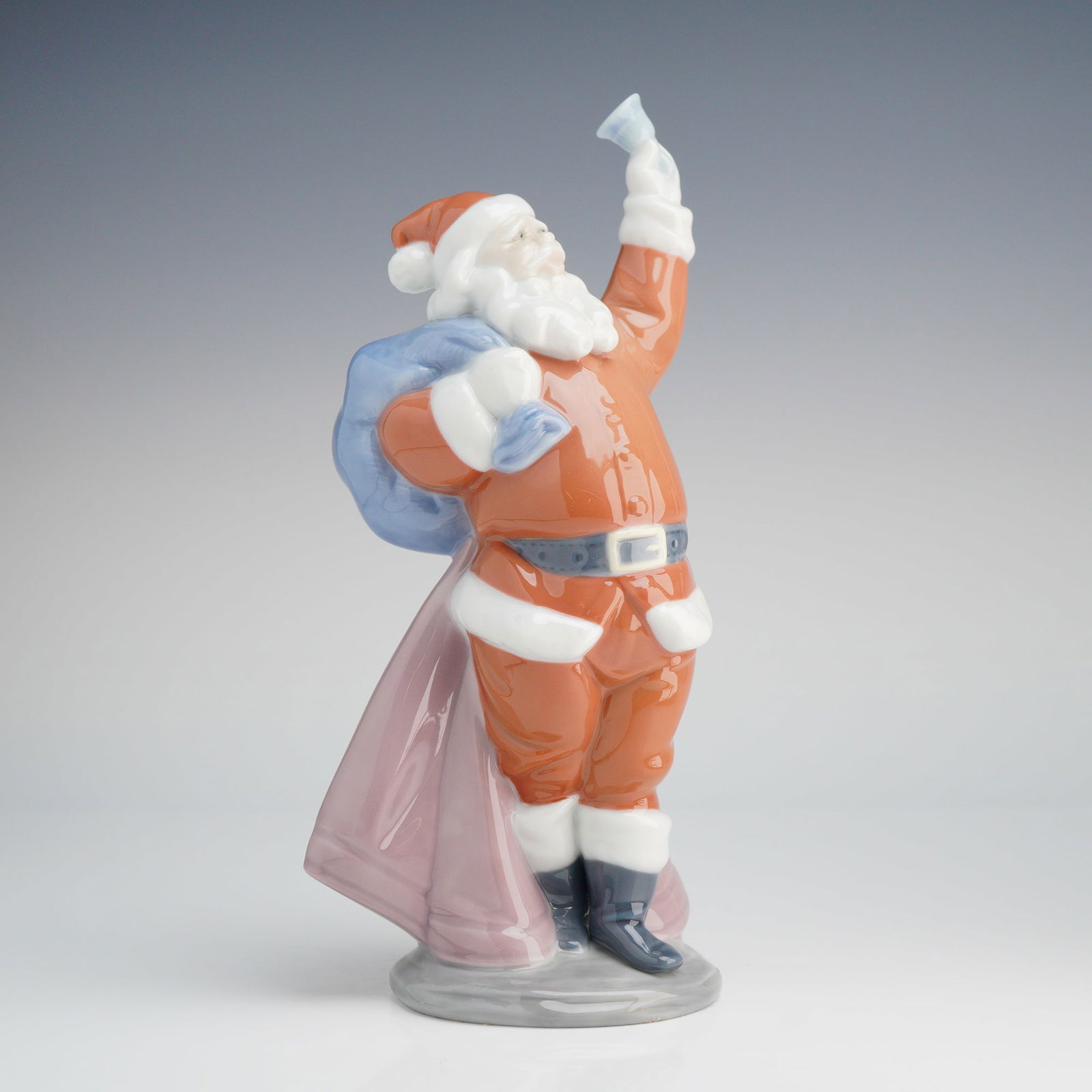 Lladro Jolly Santa Porcelain Figurine 6500: A Lladro porcelain figurine titled "Jolly Santa," model number 6500. The sculpture depicts Santa Claus dressed in a glossy orange-red suit with white trim, dark blue boots, and a belt, carrying a ligh