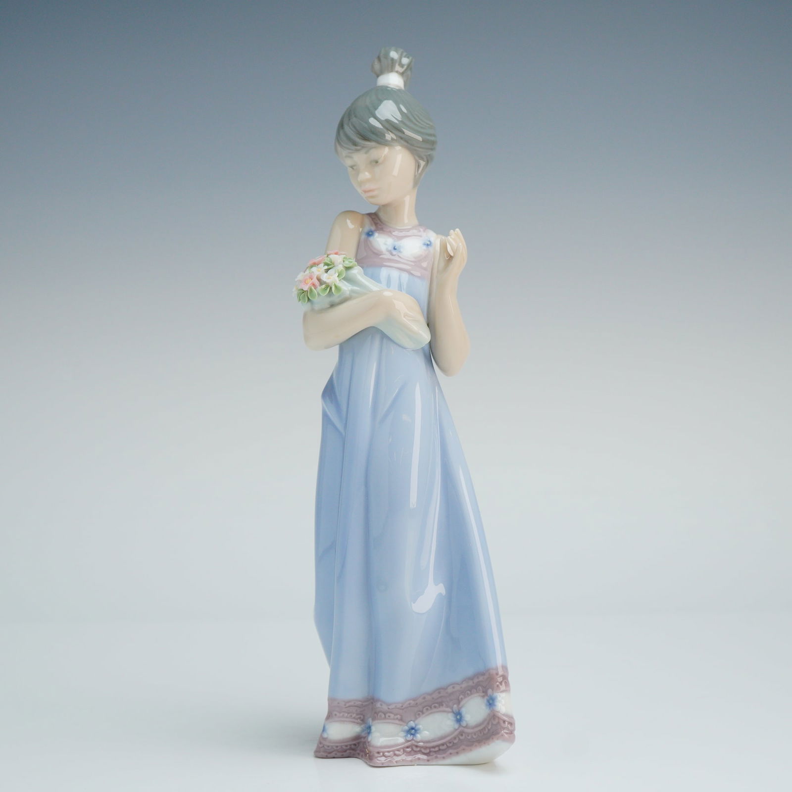 Lladro Porcelain Figurine Spring Token 01005604: The piece depicts a young woman with hair styled in a high bun, wearing a sleeveless, floor-length pale blue gown with pink and white floral embroidery at the neckline and hem. She is shown cradling a