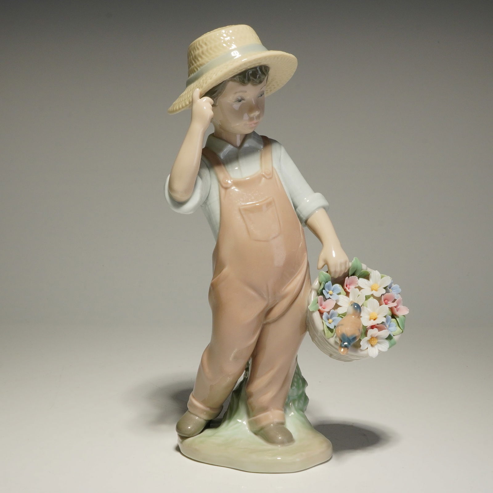 Lladro Porcelain Figurine, My Happy Friend 01006824: This Lladro depicts a young boy dressed in a straw-textured hat, a light blue long-sleeved shirt, and tan overalls. The figure is shown carrying a basket filled with various pastel-colored flowers, wi