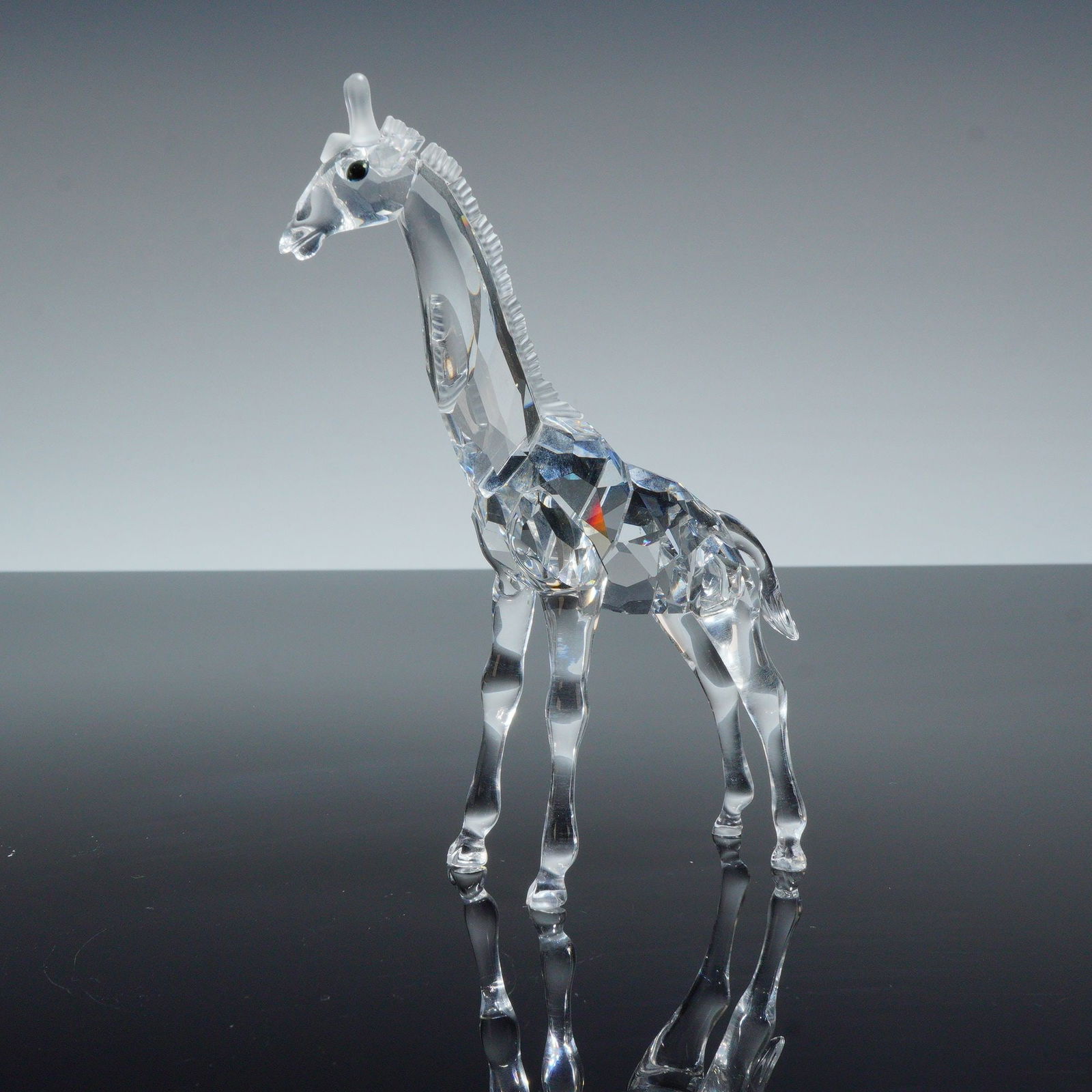 Swarovski Crystal Figurine, Baby Giraffe 236717: Crafted from faceted lead crystal. The design features a faceted torso and neck, a textured mane, and smooth, tapered legs. The head is detailed with small, dark bead eyes. The Swarovski swan maker ma