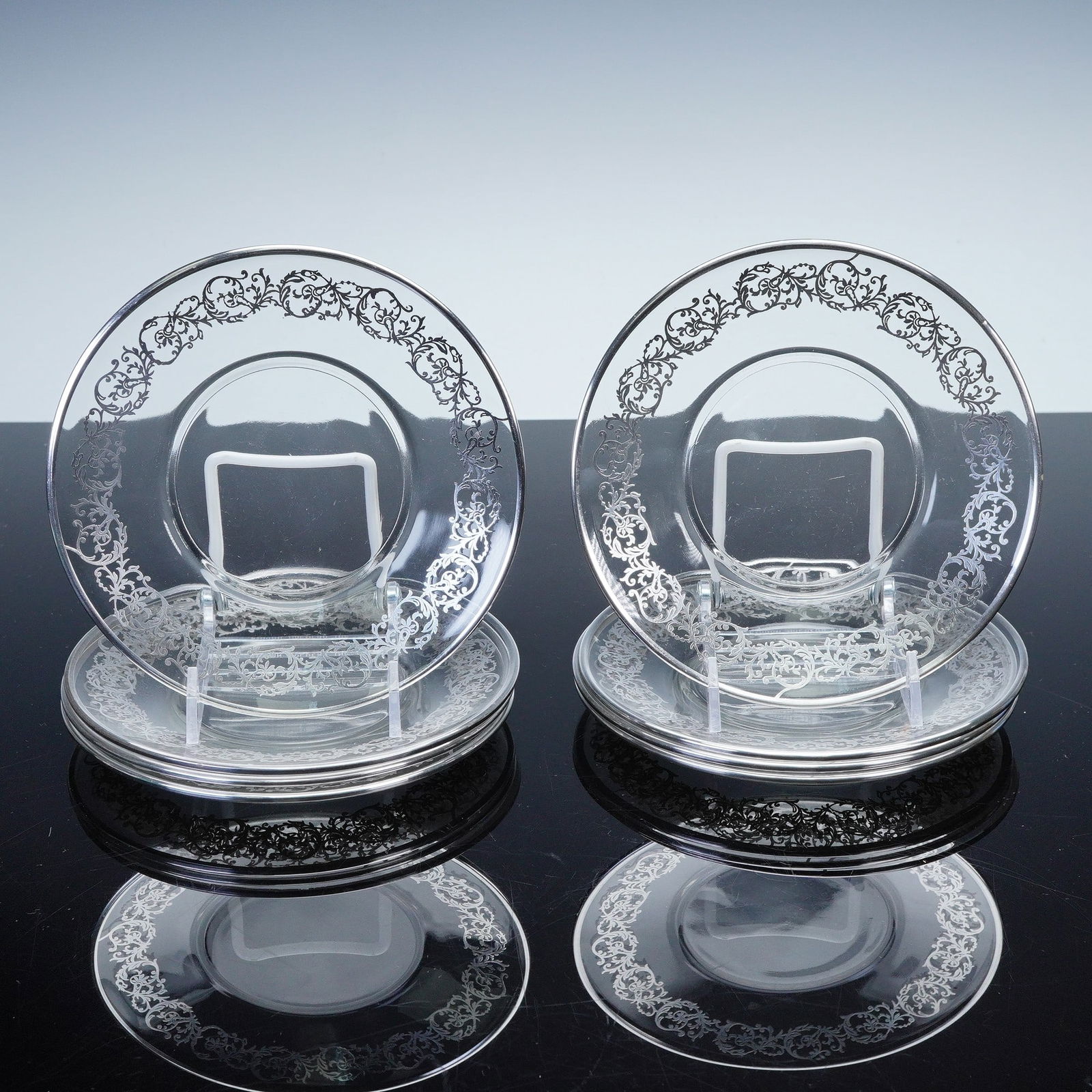 8pc Silver Toned Art Glass Plates: Set of eight clear glass plates with ornate silver toned overlay in a scrolling floral design around the borders. The decoration creates an elegant contrast against the transparent ground, adding a