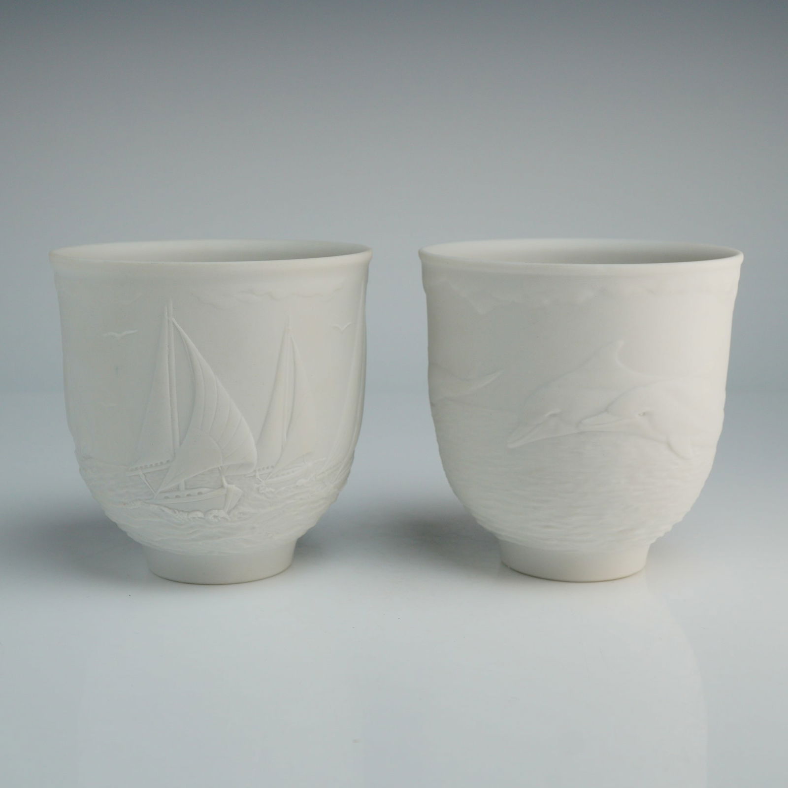 2pc Lladro White Bisque Porcelain Votive Cups: A grouping of Lladro Collectors Society white bisque porcelain candle holders featuring high-relief molded maritime scenes. The first holder, titled Sailing The Seas (01017657), depicts sailboats on t