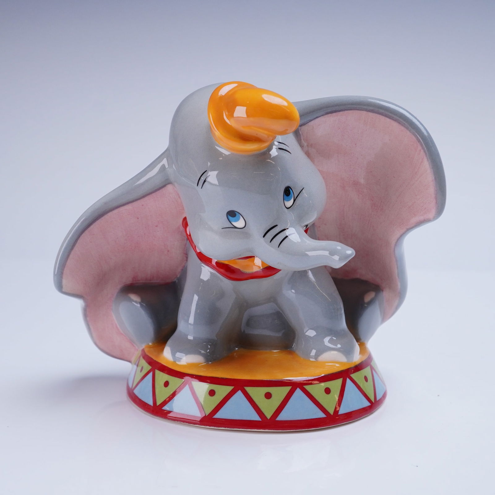 Royal Doulton Disney Showcase Collection Figurine Dumbo FC 3: A glazed ceramic figurine depicting the Walt Disney character Dumbo, seated on a brightly colored circus pedestal decorated with geometric patterns. The piece is rendered in high-gloss gray, pink, ora