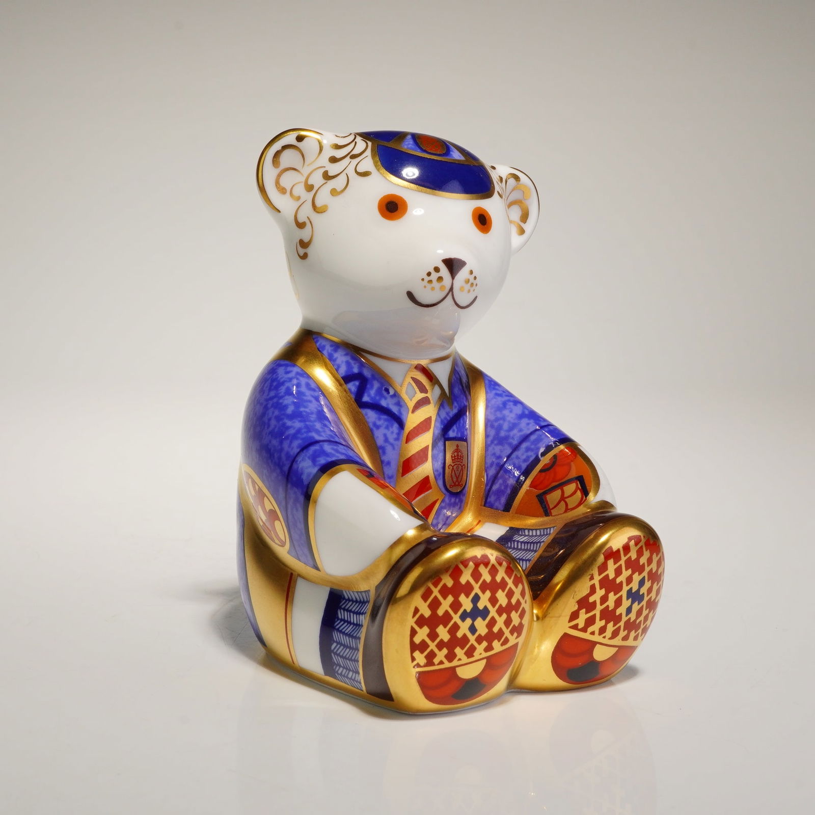 Royal Crown Derby Schoolboy Teddy Paperweight: A Royal Crown Derby fine bone china paperweight titled Schoolboy Teddy. The figurine depicts a seated bear in collegiate attire, featuring a textured cobalt blue blazer with a crest, a red and gold st
