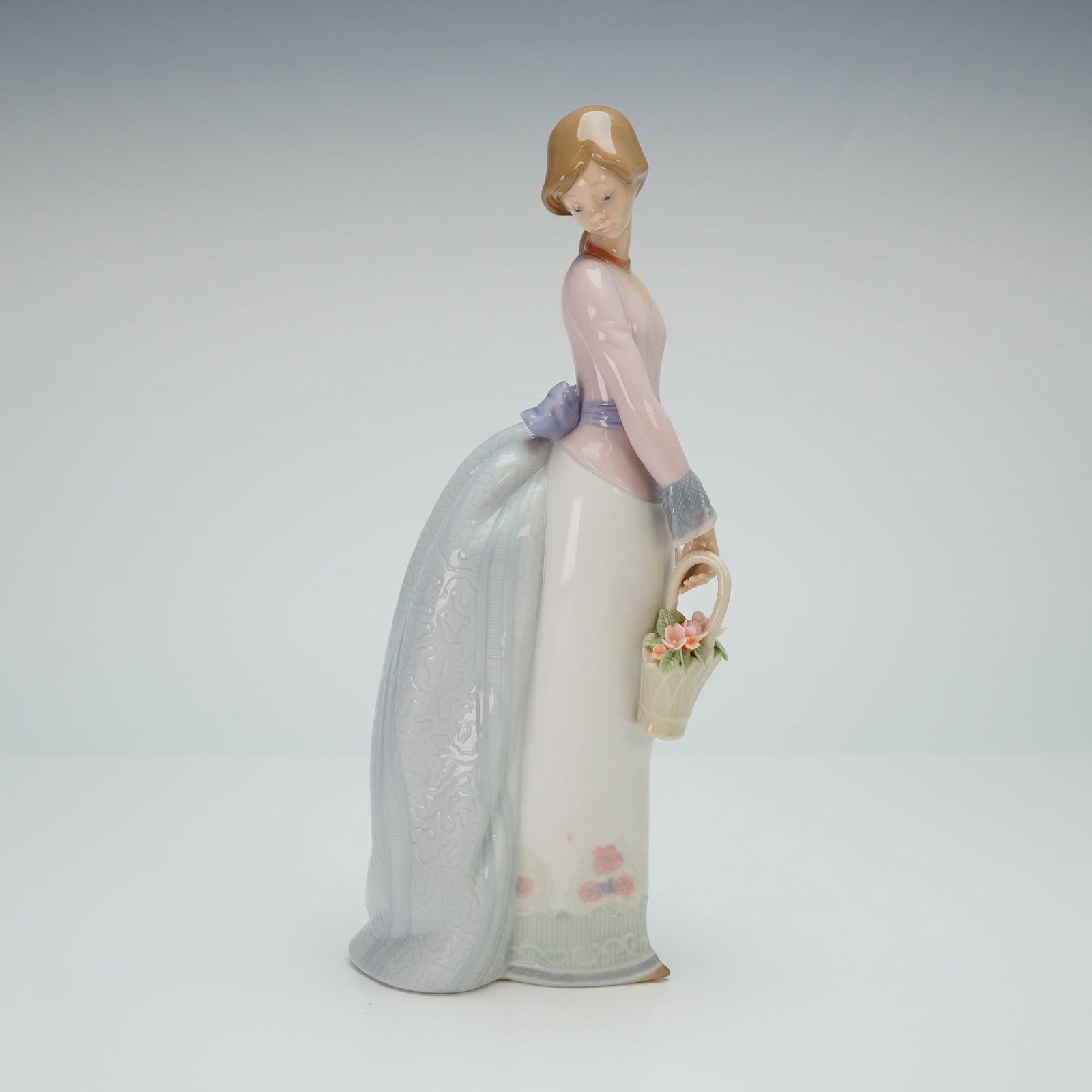 Lladro Porcelain Figurine Basket of Love 01007622: Lladro originally issued for the Lladro Collectors Society. This glazed porcelain figure depicts a young woman in a long, pale pink and white gown featuring a decorative blue bustle and a purple bow a