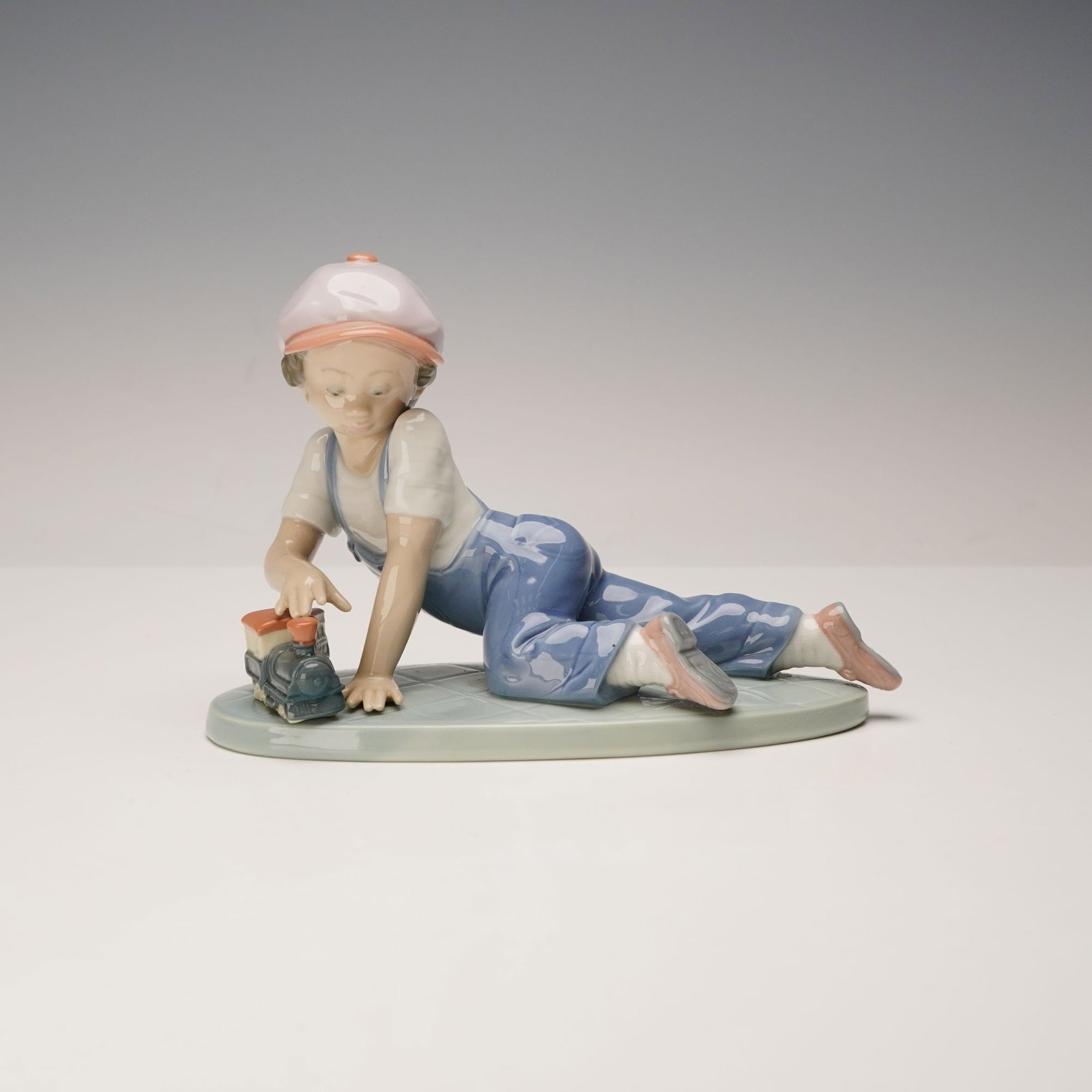 Lladro Porcelain Figurine, All Aboard 1007619: Issued for the Collectors Society in 1993. The glazed figurine depicts a young boy in a white shirt, blue overalls, and a pink baseball cap, lying on his stomach and playing with a two-car toy train o