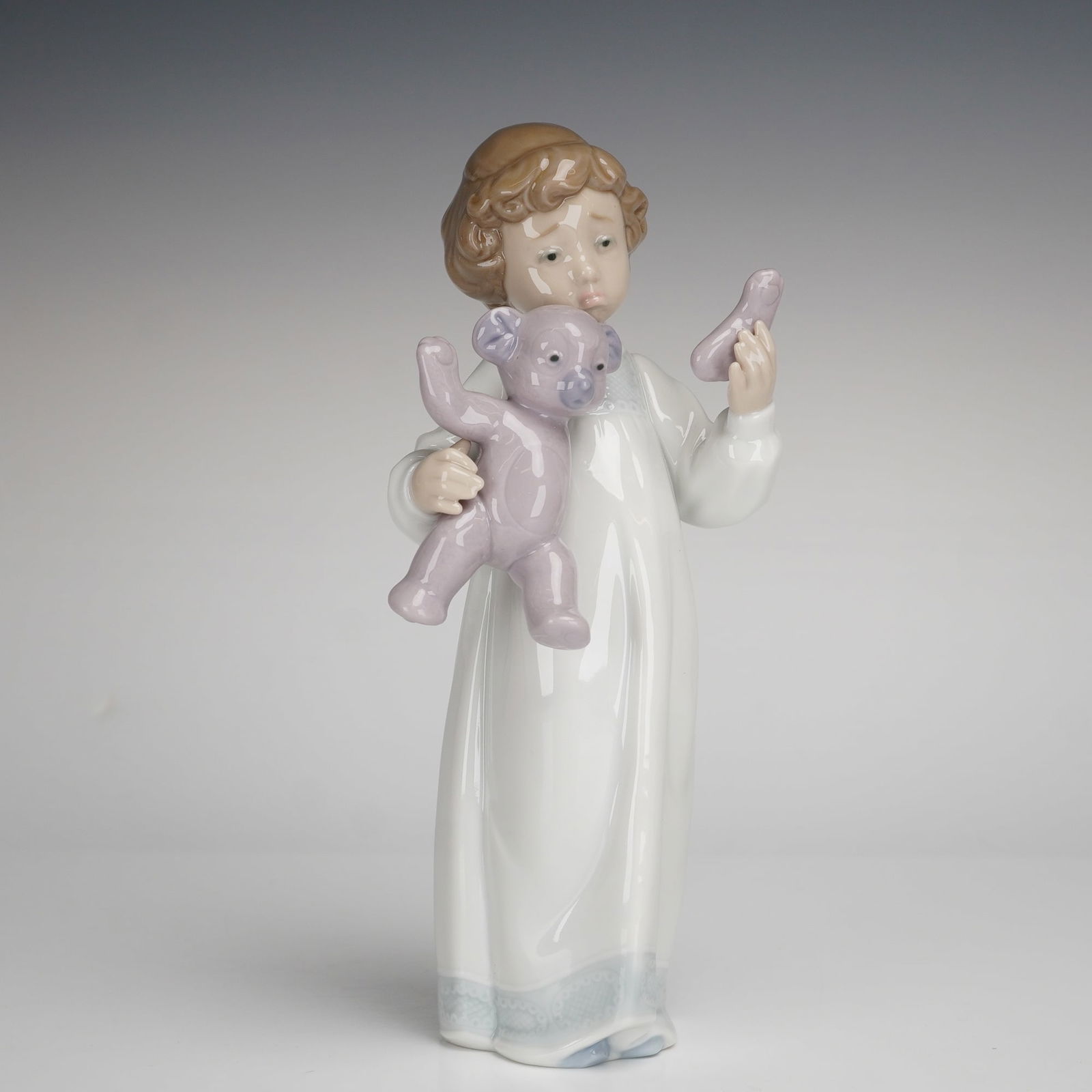 Nao by Lladro Porcelain Figurine, Poor Teddy!: A Nao by Lladro glazed porcelain figurine titled Poor Teddy!. The sculpture depicts a young child with short brown hair dressed in a long white nightgown with a pale blue patterned hem. The figure is