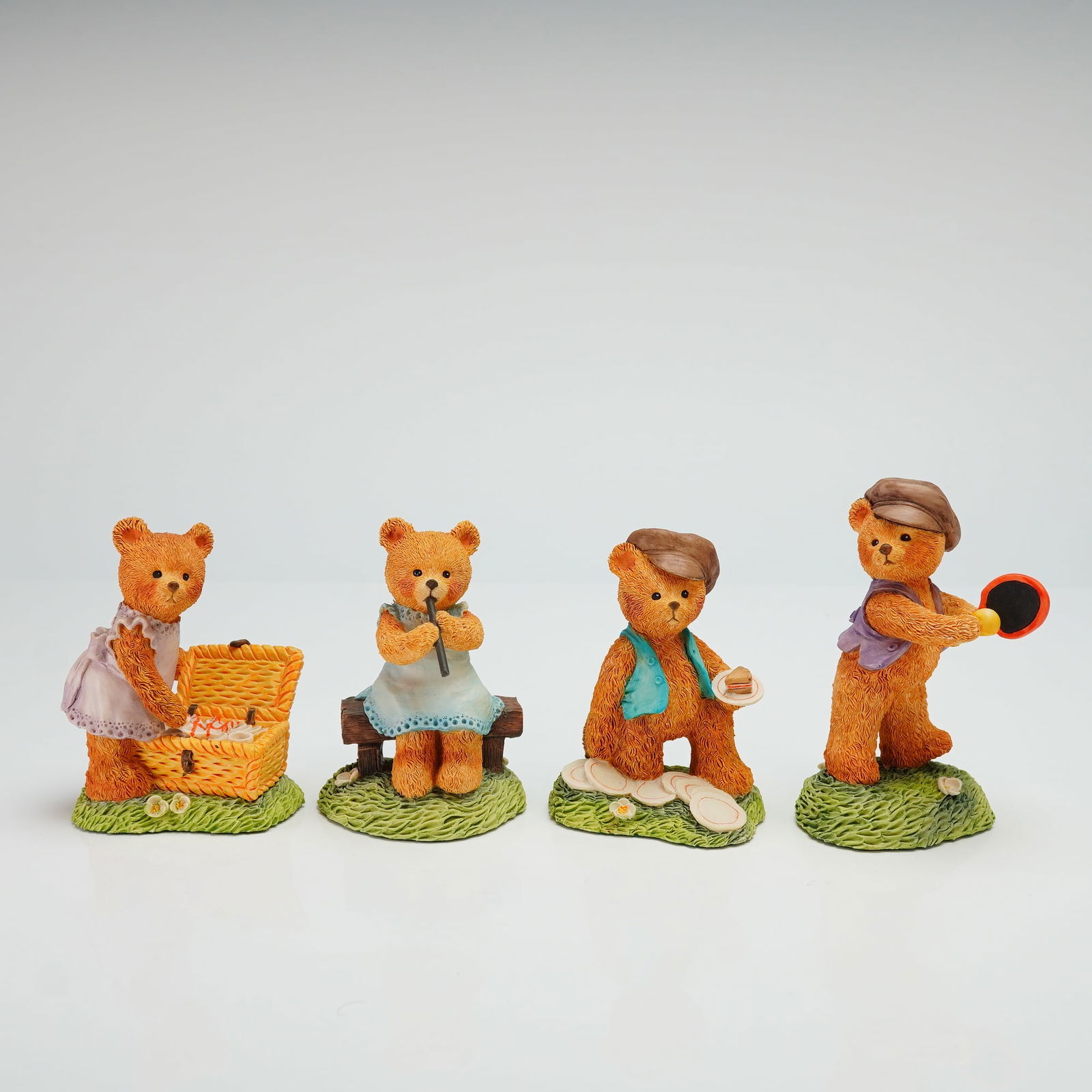 Group of Four Beswick Bear Figurines: A group of anthropomorphic teddy bear figurines constructed from cold-cast resin with a polychrome painted finish. Each figure is set upon a textured base modeled as grass. The group features four dis