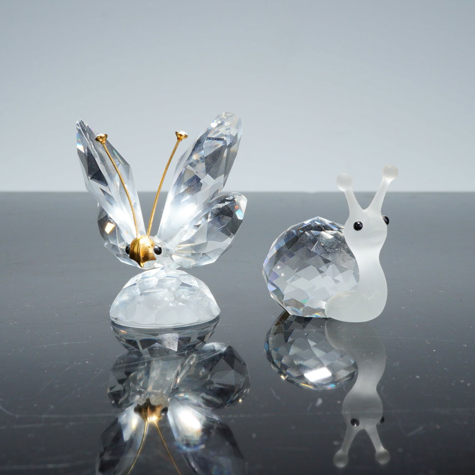 Two Swarovski Crystal Figurines, Butterfly with Gold Antennae and Snail: This lot features two Swarovski crystal figurines, consisting of a butterfly with gold antennae and a snail. The butterfly is composed of multiple faceted clear crystal components forming the wings an
