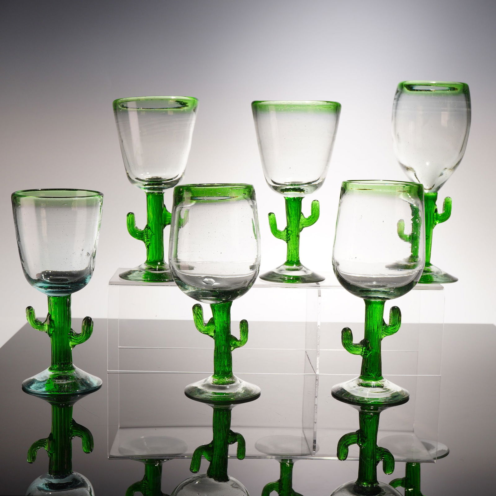 6pc Hand-Blown Cactus Stem Glasses: A collection of six artisanal glasses, each featuring clear glass bowls accented with applied emerald green rims. The figural stems are expertly modeled in the form of cacti in a vibrant green glass,