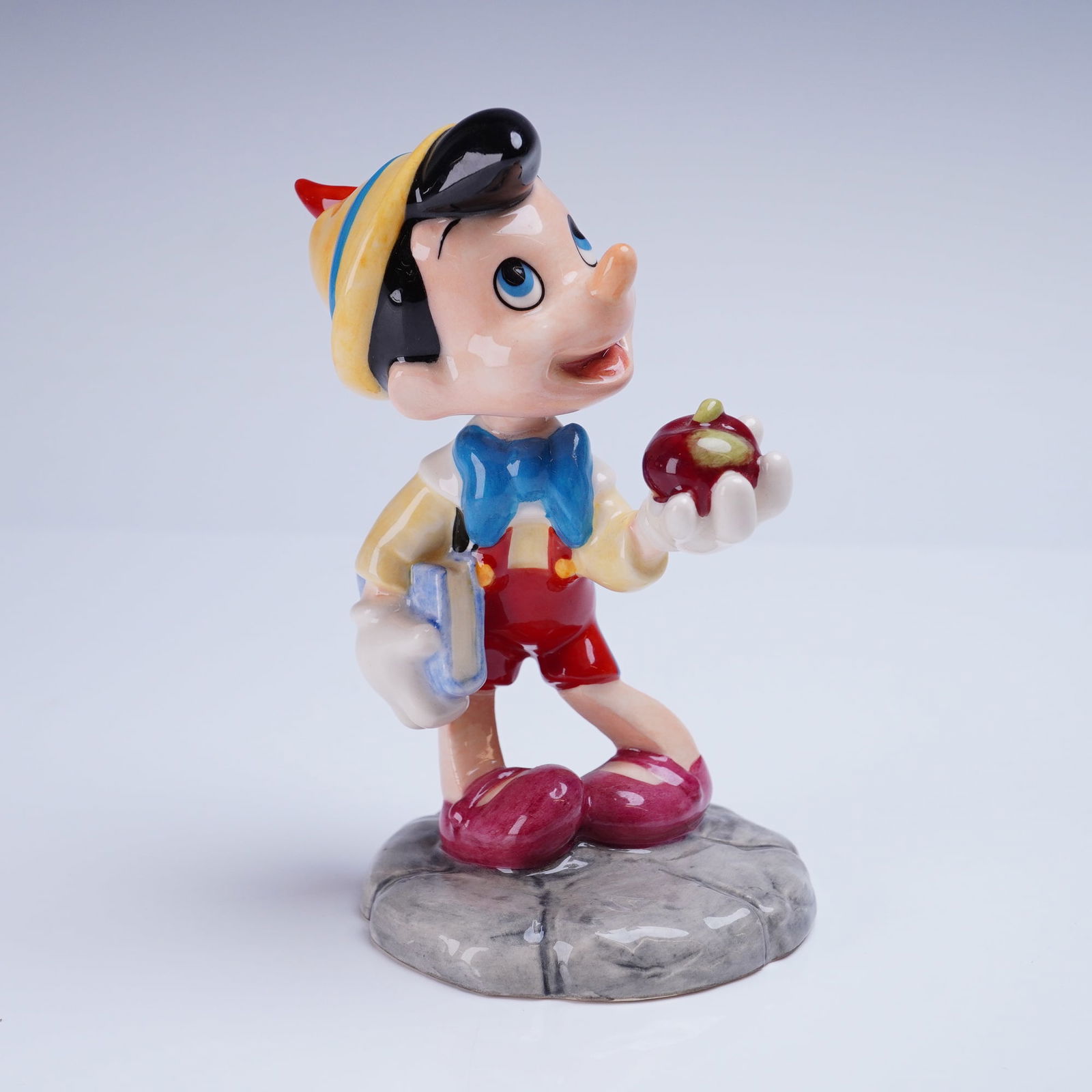 Royal Doulton Disney Showcase Collection Pinocchio Figurine FC 4: A glazed ceramic figurine from the Royal Doulton Film Classics Collection, titled "Pinocchio". The figurine depicts the character Pinocchio standing on a textured grey base, holding a red apple in his
