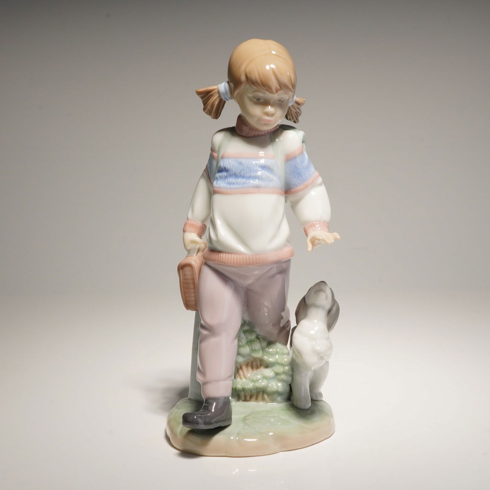 Lladro Porcelain Figurine Thursday's Child 01006018: This Lladro glazed porcelain figurine depicts a young schoolgirl with blonde pigtails walking alongside a small dog near a milestone post. The figure is detailed with a white and blue textured sweater