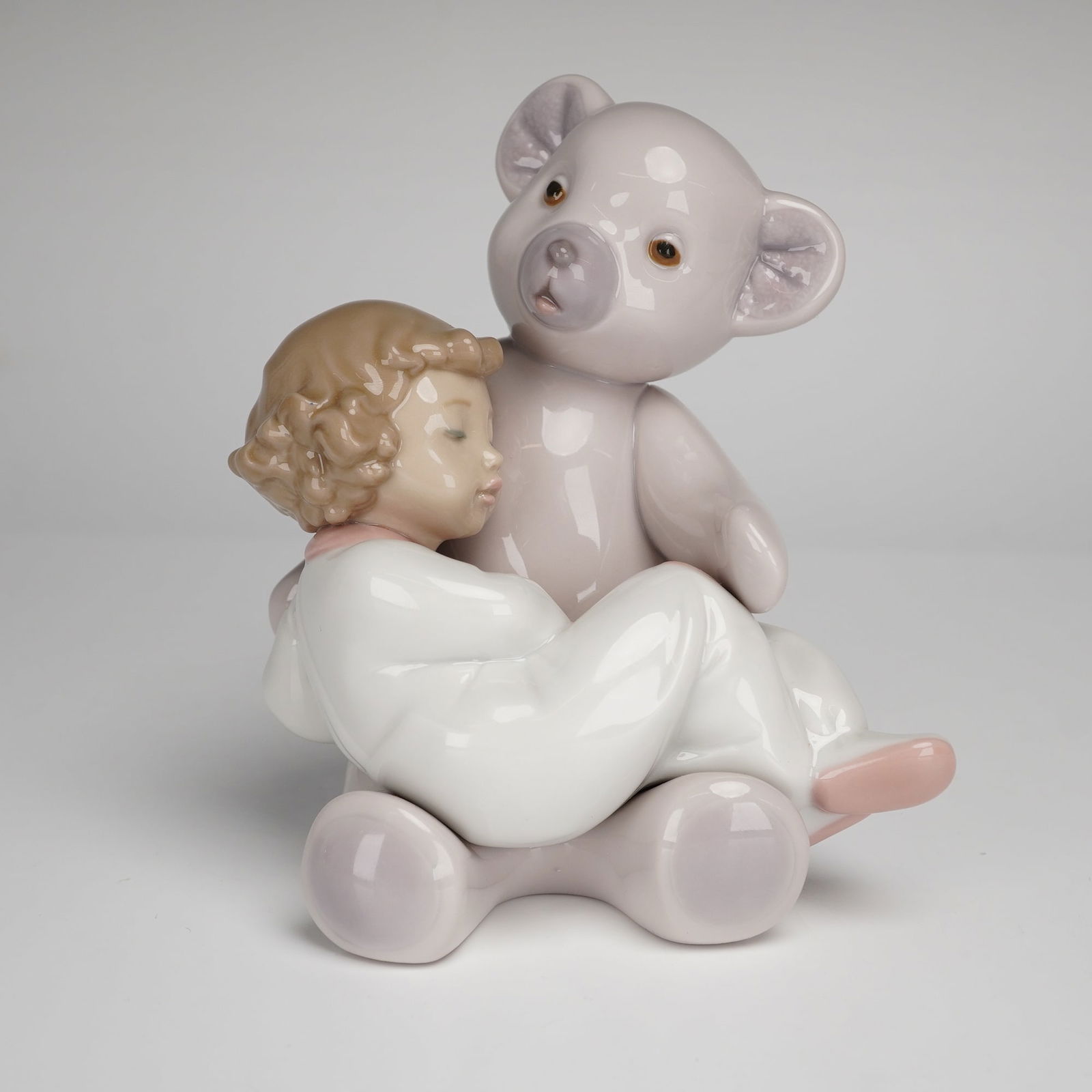 Nao by Lladro Porcelain Figurine, Big Bear Hug: Nao by Lladro porcelain figurine titled Big Bear Hug. This glazed porcelain piece depicts a young child in white pajamas sleeping while leaning against a large light gray teddy bear. The figurine feat