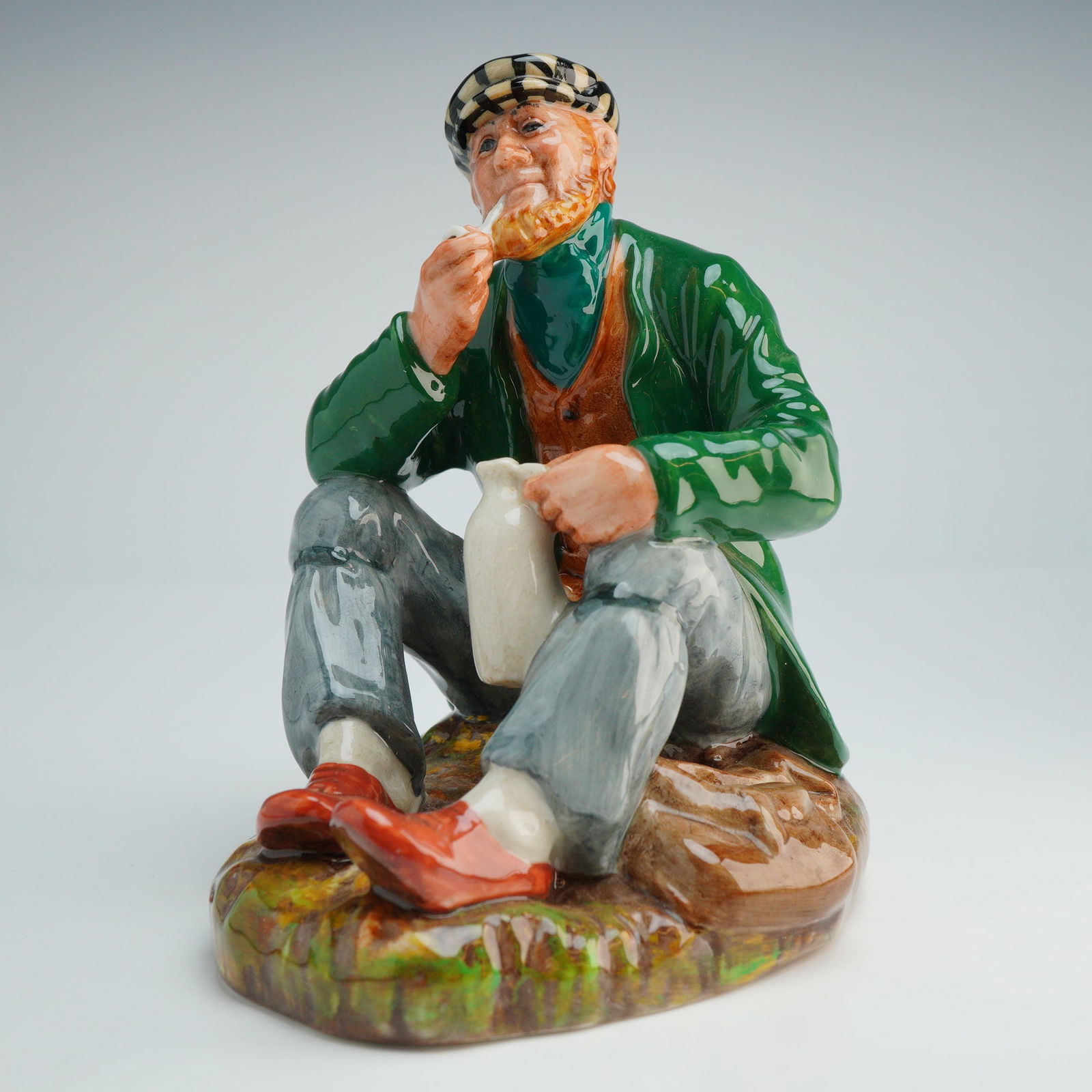 Royal Doulton Figurine The Wayfarer HN2362: This glazed ceramic piece features a high-gloss finish and depicts a seated, bearded man. The figure is dressed in a green overcoat, orange waistcoat, grey trousers, and a black and white checkered ca