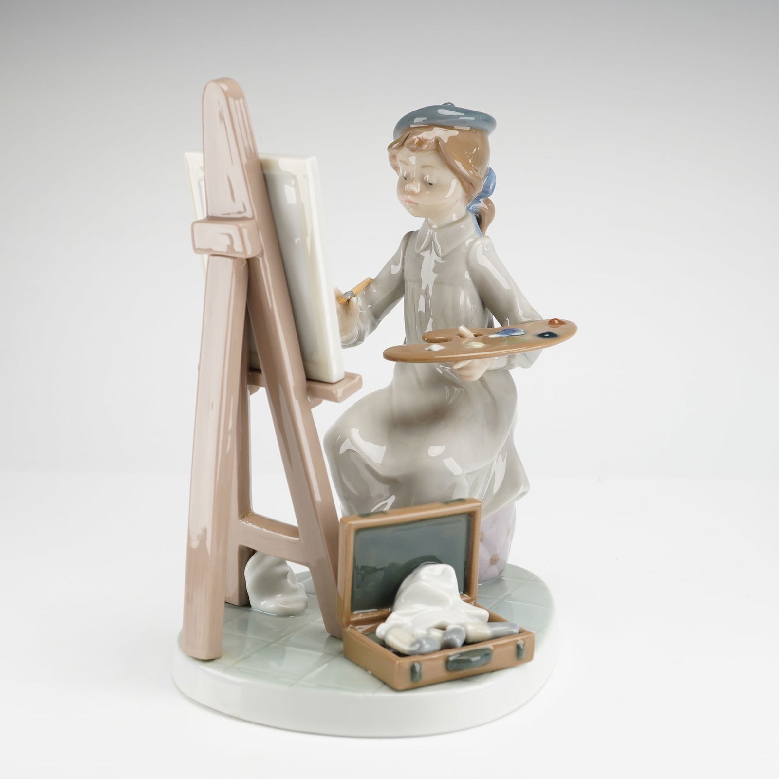Lladro Still Life Artist Porcelain Figurine 01005363: This Lladro Still Life Artist (01005363) porcelain figurine depicts a young girl seated on a tufted ottoman painting a floral scene on a canvas supported by an easel. The figure is constructed of hand