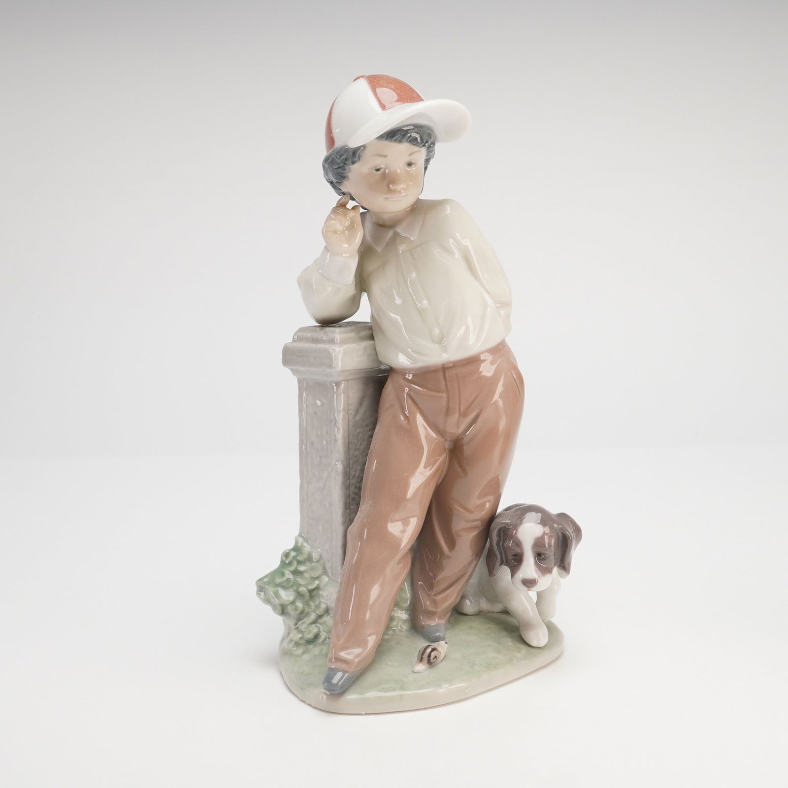 Lladro Best Foot Forward Porcelain Figurine, No. 5738: A fine Lladro glossy porcelain figurine titled Best Foot Forward, model number 5738. This charming study features a young boy in a relaxed pose, leaning against a stone-style pedestal. He is dressed i