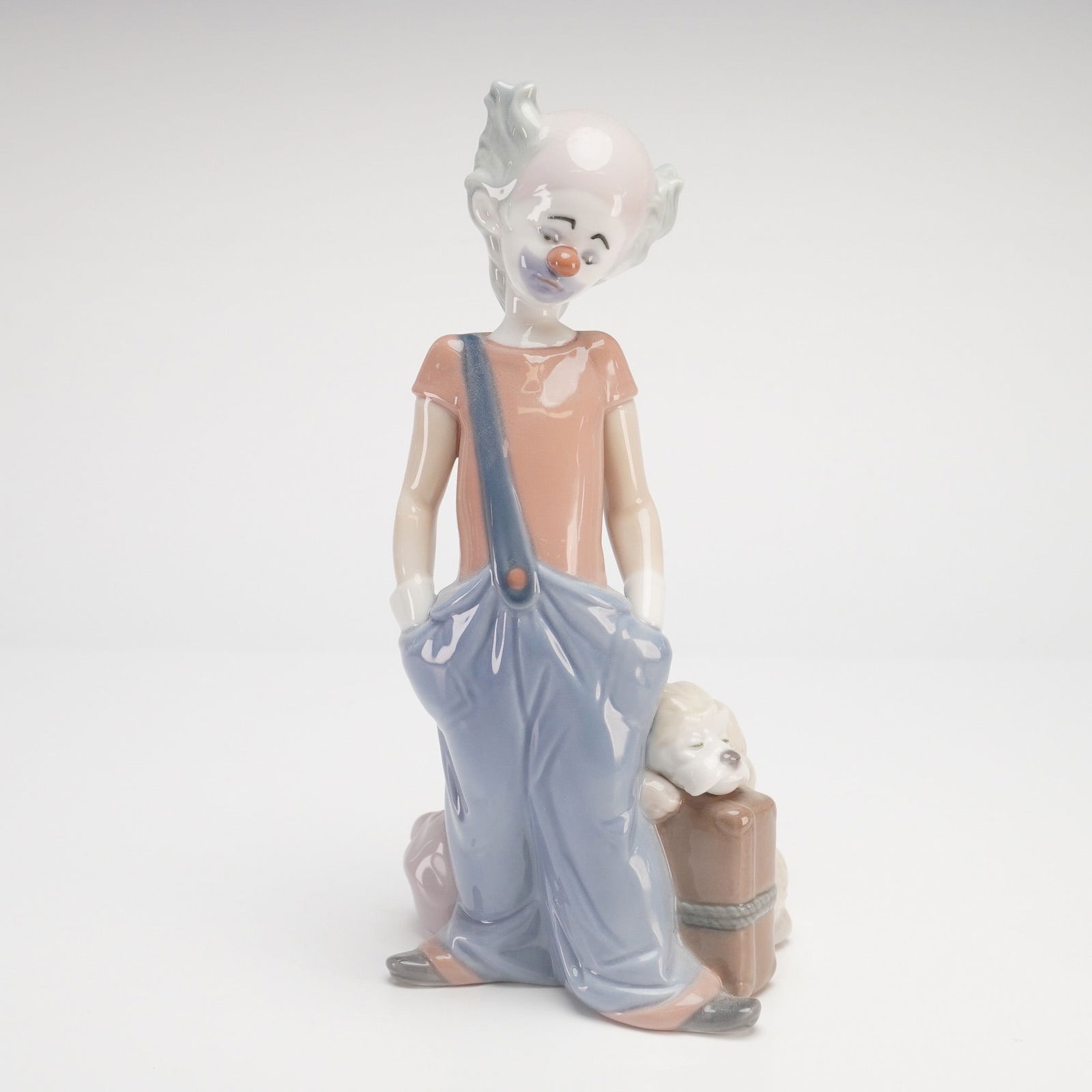 Lladro Destination Big Top Clown Figurine, Model 6245: This Lladro glossy porcelain figurine, titled Destination Big Top (model 6245), depicts a melancholic circus clown in a whimsical, character-driven pose. The figure is shown wearing oversized, sagging