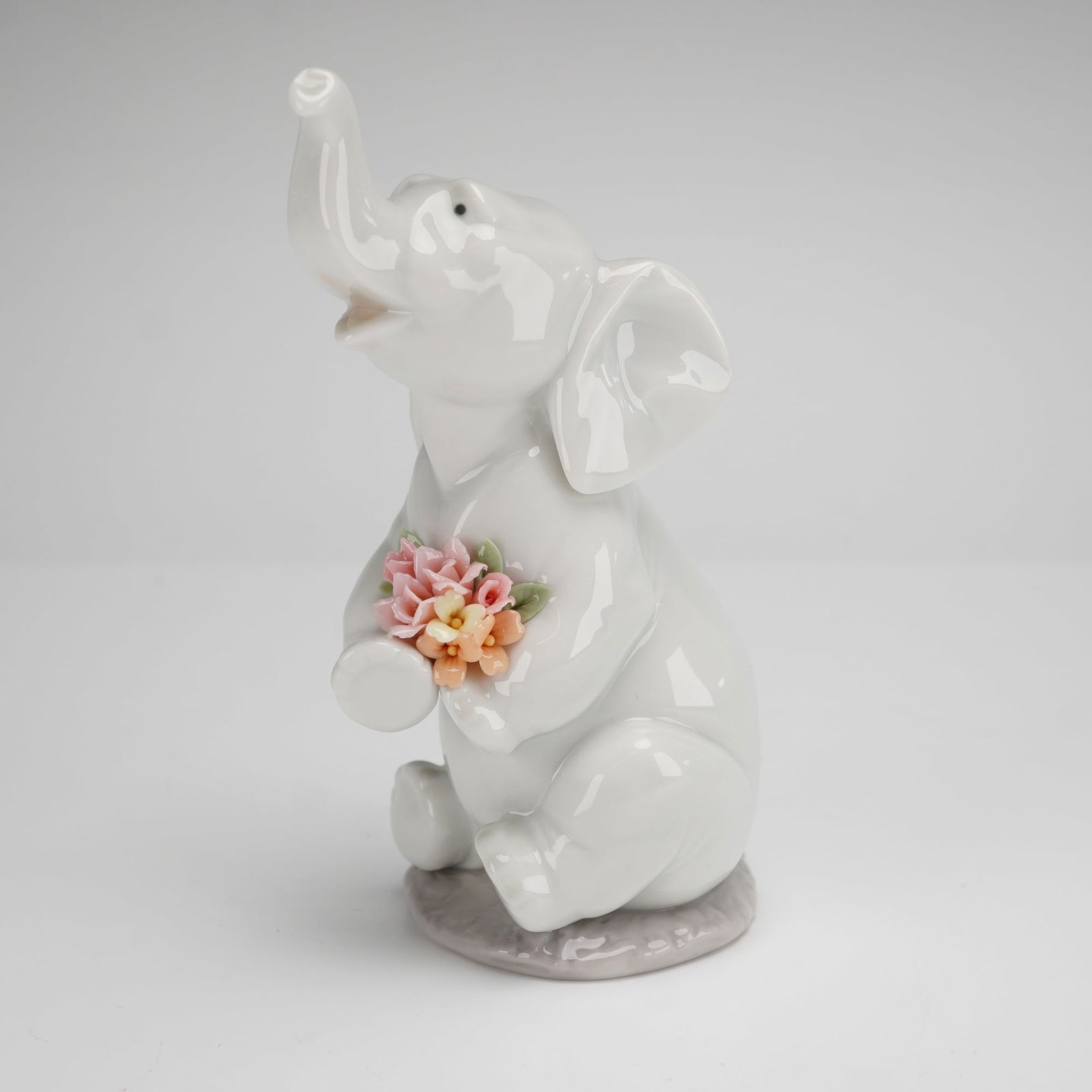 Lladro Lucky in Love Porcelain Figurine 6462: This Lladro porcelain figurine, titled Lucky in Love (model 1006462), depicts a seated baby elephant with its trunk raised. The figure holds a bouquet of hand-formed flowers in pastel shades of pink,