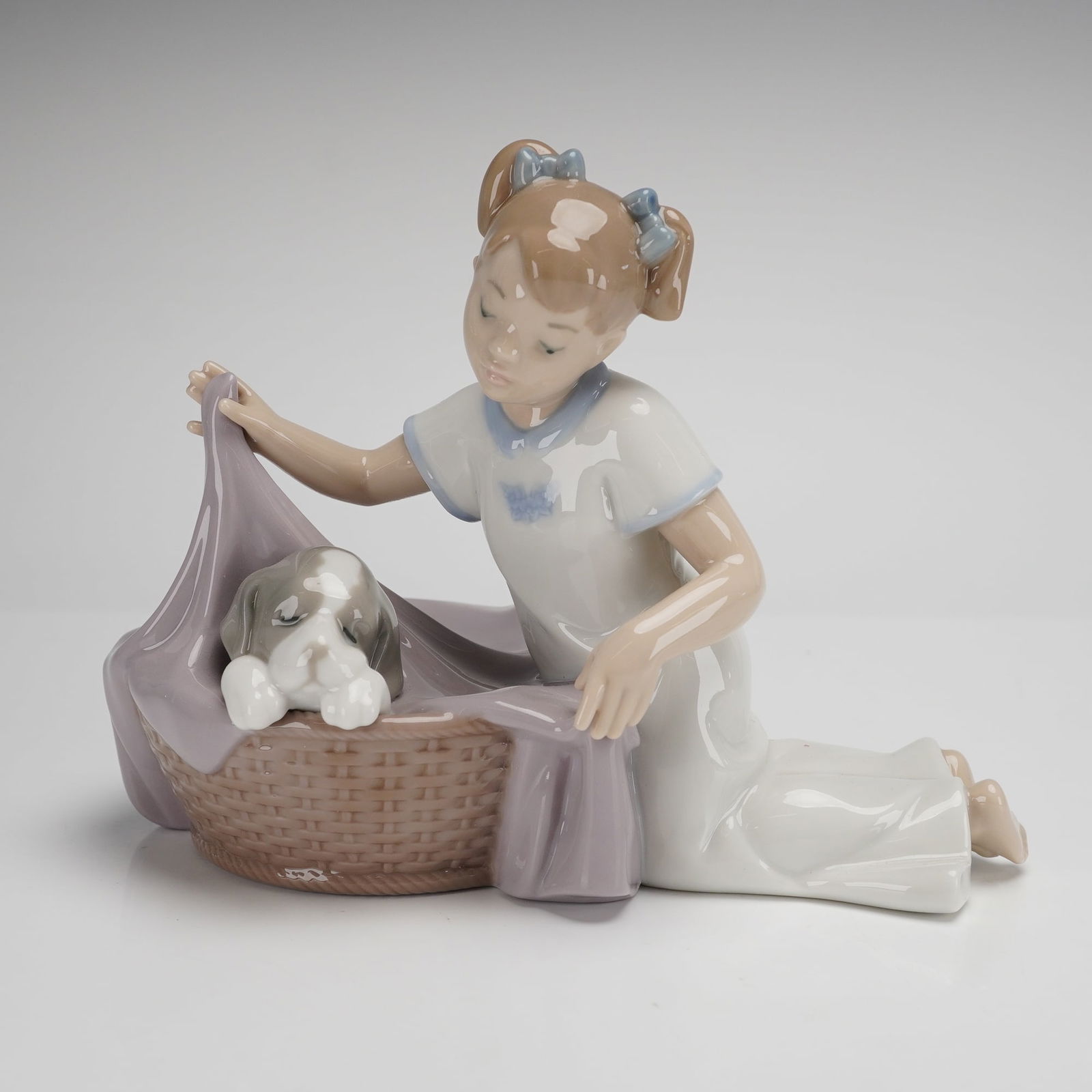 NAO by Lladro Porcelain Figurine, It's Time to Sleep: A glazed porcelain figurine by NAO, a division of Lladro, titled It's Time to Sleep. The piece depicts a young girl with her hair in pigtails wearing a white nightgown, kneeling beside a woven-texture
