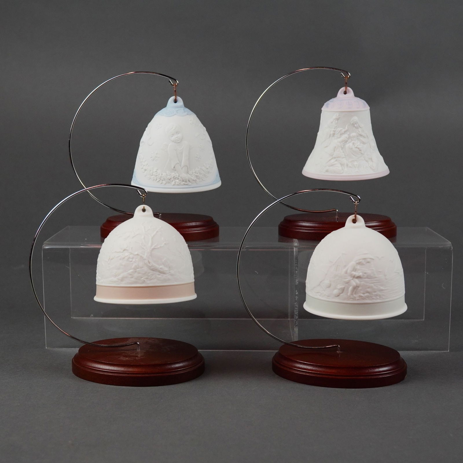 Lladro Collectors Society Bisque Porcelain Bell Grouping with Stands: A four-piece Lladro bisque porcelain bell grouping featuring the annual Collectors Society series from 1990 through 1993. Each bell showcases a seasonal theme in bas-relief: the 1990 Spring Bell with