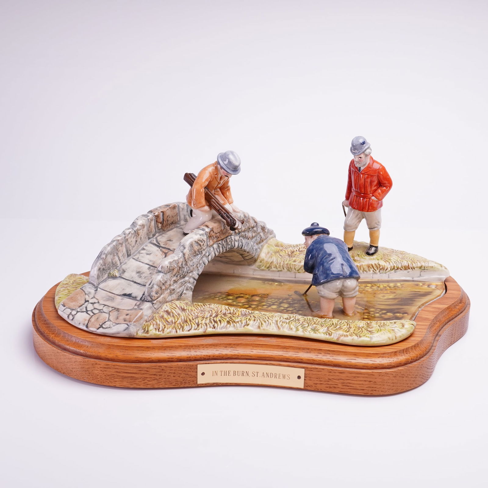 Royal Doulton Figurine, In the Burn, St. Andrews D6890: This hand-painted porcelain work depicts a historical golfing scene with three figures in traditional attire navigating a stone bridge and stream. The grouping is mounted on a contoured wooden base fe