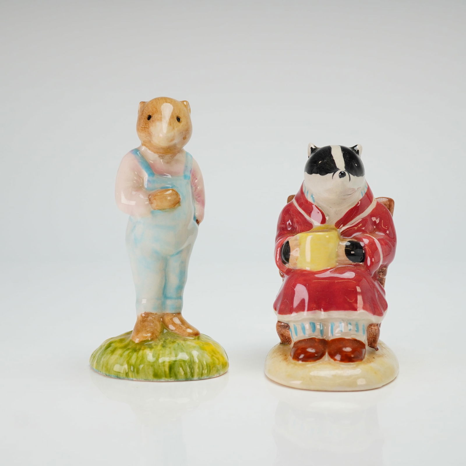 Two Royal Albert Beatrix Potter Figurines, Ratty and Badger: The figure of Ratty (AW3) is depicted standing in blue overalls on a grassy base. The figure of Badger (AW2) is depicted seated in a wooden chair wearing a red robe and holding a yellow mug. Both piec