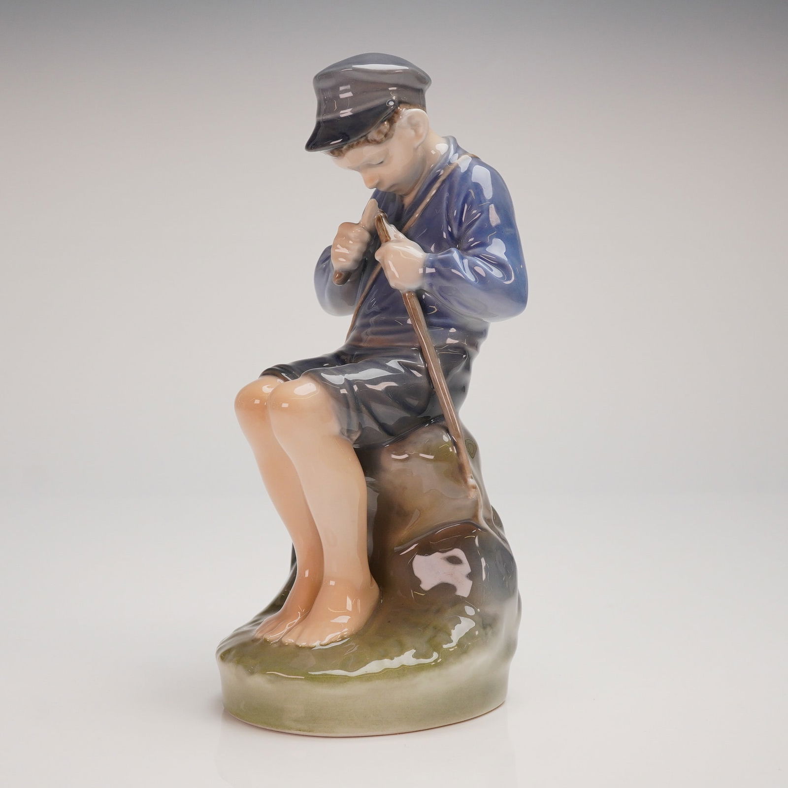 Royal Copenhagen Boy Whittling a Stick Porcelain Figurine Model 905: A single glazed porcelain figurine by Royal Copenhagen, model number 905, depicting a young boy seated barefoot upon a naturalistic rock formation while contemplating a slender stick or staff held in