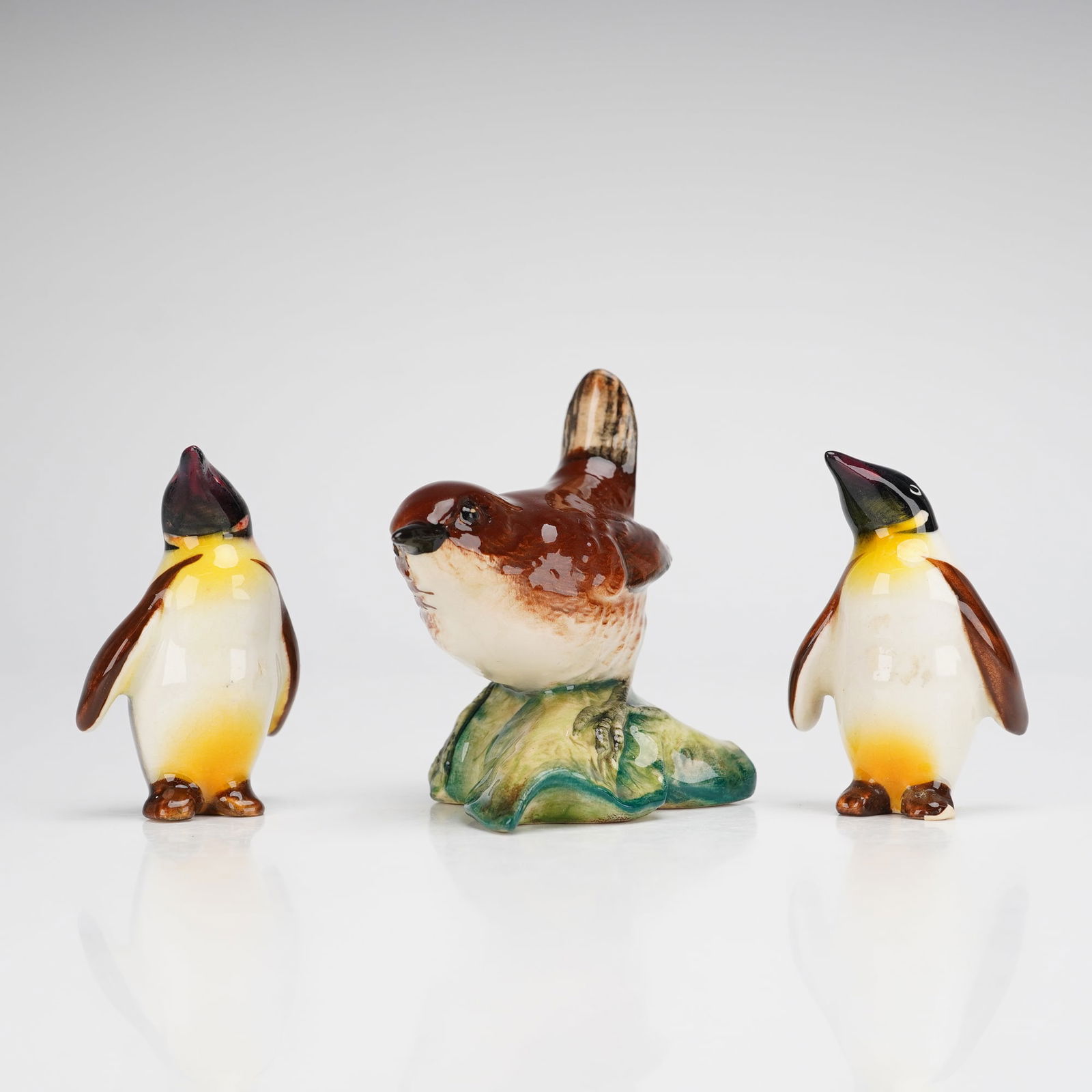 Three Beswick Bird Figurines, Penguins and Wren: A trio of Beswick glazed ceramic bird figurines, including two penguins and a wren. The wren is naturalistically modeled in shades of brown and white, perched on a green and brown base. The underside