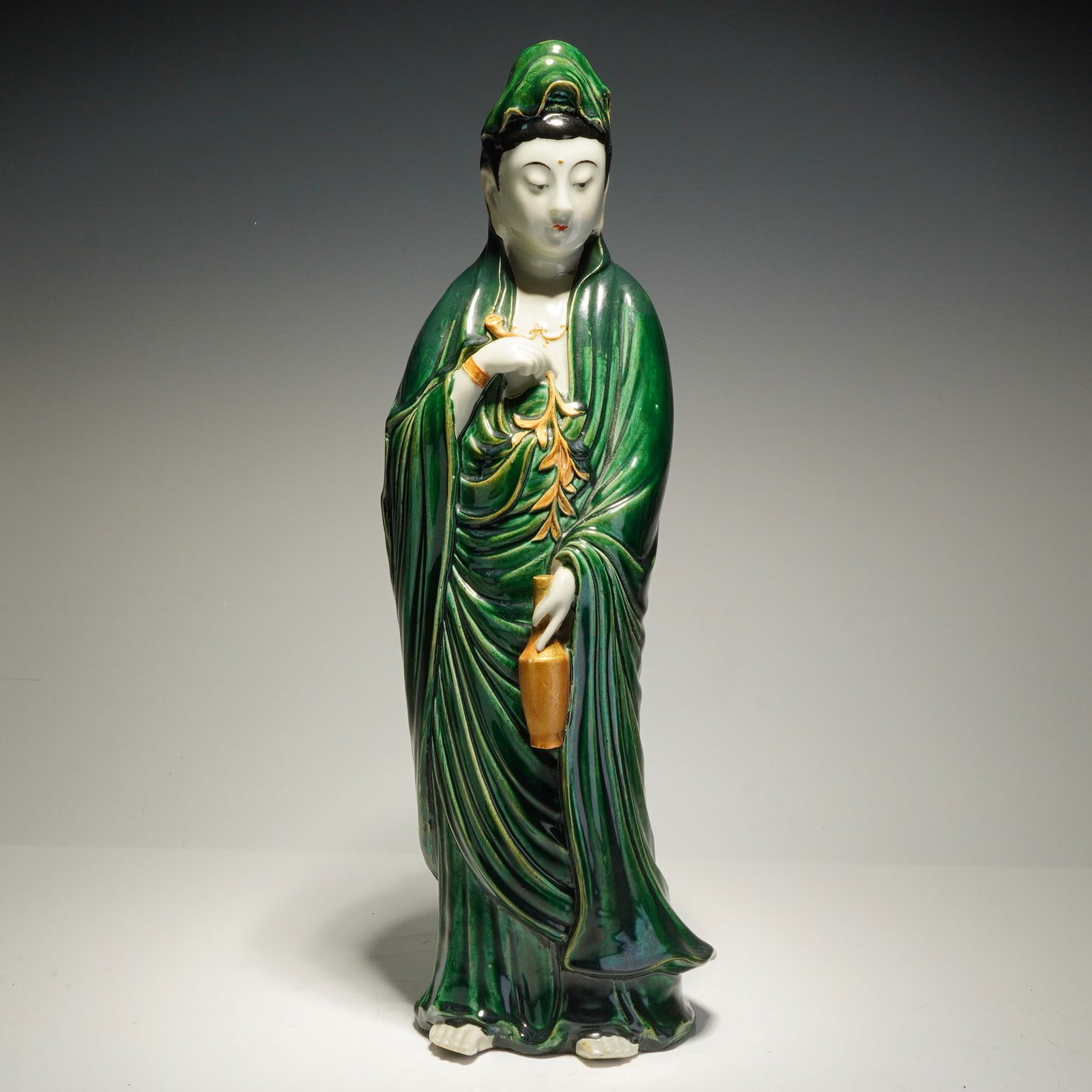 Chinese Green Glazed Ceramic Guanyin Sculpture: This Chinese ceramic sculpture depicts a standing Guanyin. The figure is rendered in hooded, flowing robes finished in an emerald green glaze. The face, hands, and feet are finished in a white glaze,