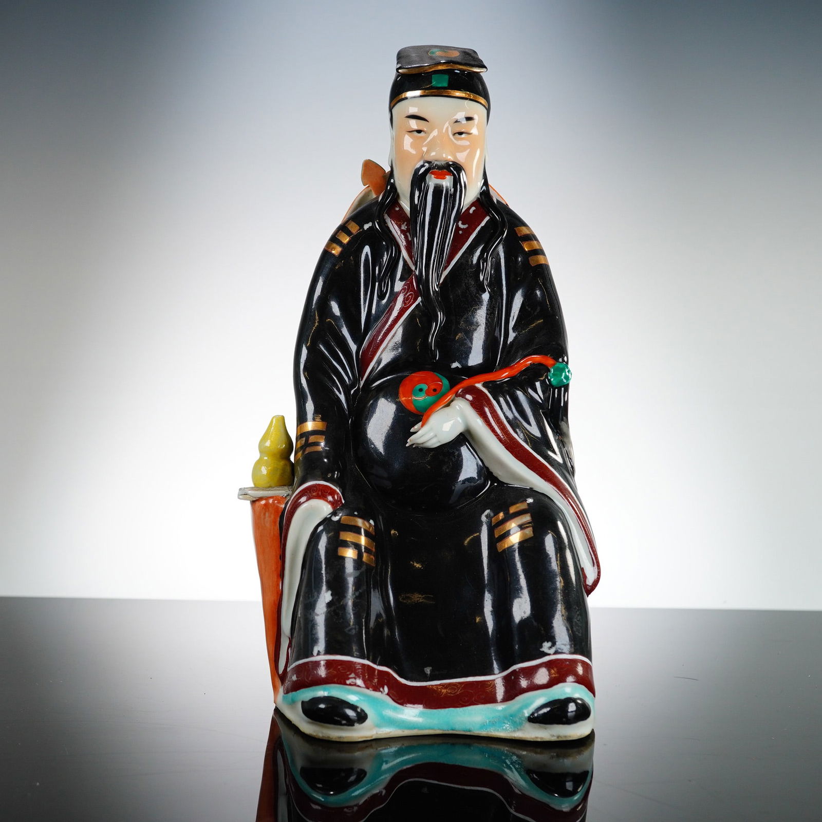 Chinese Ceramic Celestial Deity Figurine, Lu Xing: Lu Xing, the Star of Prosperity, rendered in hand painted and gilded polychrome glazed porcelain. The figure is modeled in a seated posture on an orange-red textured base, wearing an official's cap an