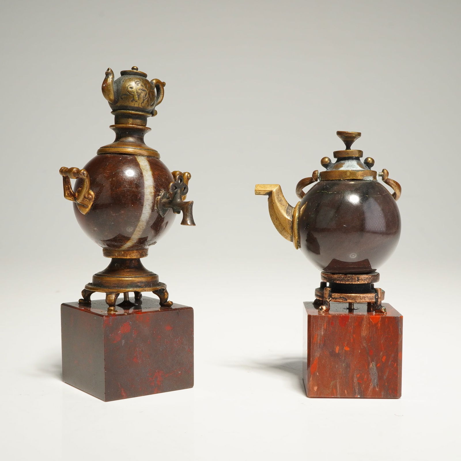 2pc Brass and Stone Teapot and Samovar Figurines: A grouping of miniature decorative objects comprising a samovar and a teapot. Each vessel features a spherical body of polished dark reddish-brown stone. The pieces are adorned with cast brass mounts