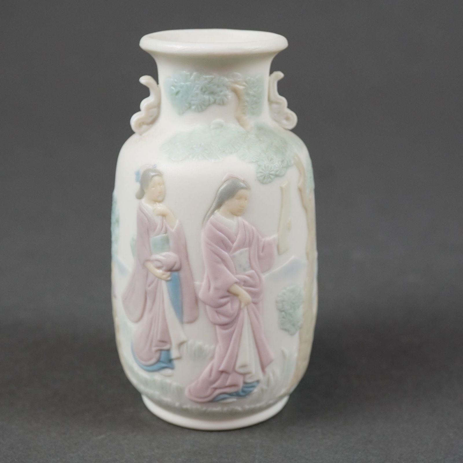 Circa 1984 Lladro Matte Porcelain Decorated Vase: A small ovoid matte porcelain vase featuring a flared rim and twin scroll-form handles. The body is decorated in low relief with hand-tinted pastel colors in soft greens, pinks, and blues. The front