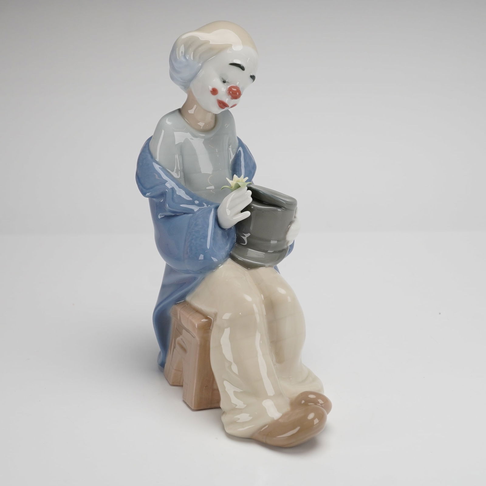 Nao by Lladro Porcelain Figurine, Little Clown in Love: This Nao by Lladro glazed porcelain figurine is titled Little Clown in Love. Hand-made in Spain, the figure depicts a seated clown with a white-painted face and red accents. The clown is dressed in a