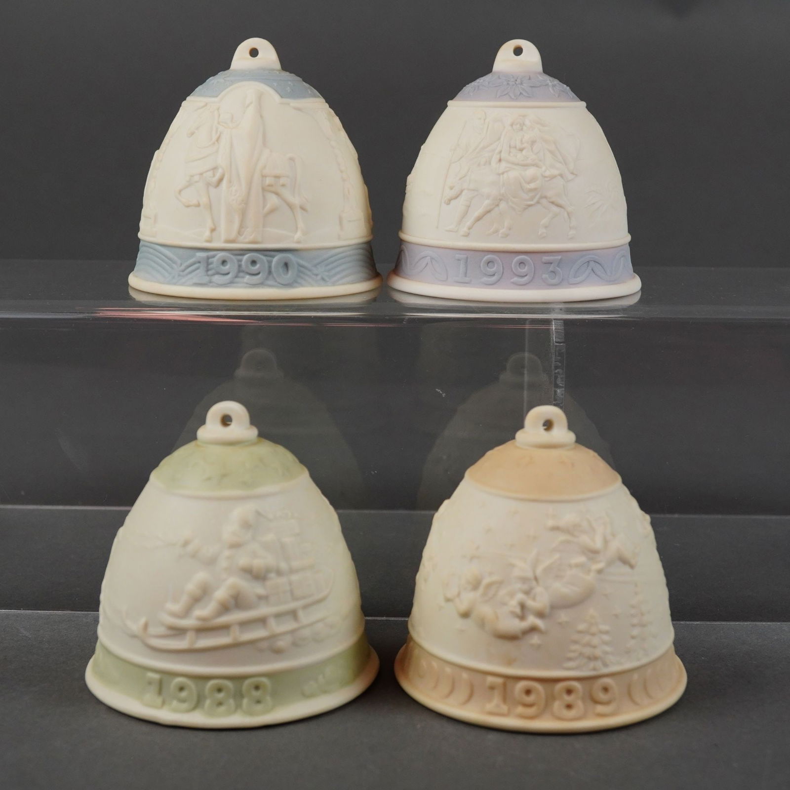 Lladro Annual Christmas Bell Grouping: A 4pc Lladro bell grouping of annual Christmas ornaments crafted in matte bisque porcelain. This collection includes the 1988 bell with green bands featuring a sledding scene; the 1989 bell with tan b