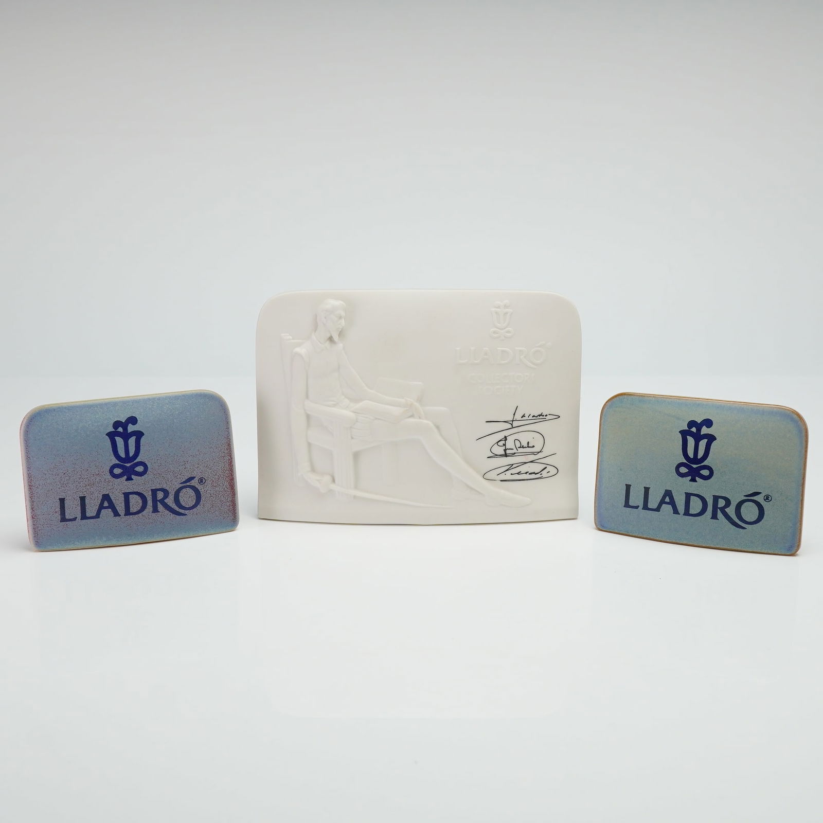 3pc Lladro Collectors Society Porcelain Plaques: This collection consists of three Lladro porcelain plaques, featuring a central Lladro Collectors Society commemorative item. The largest item is a matte white bisque porcelain plaque with a
