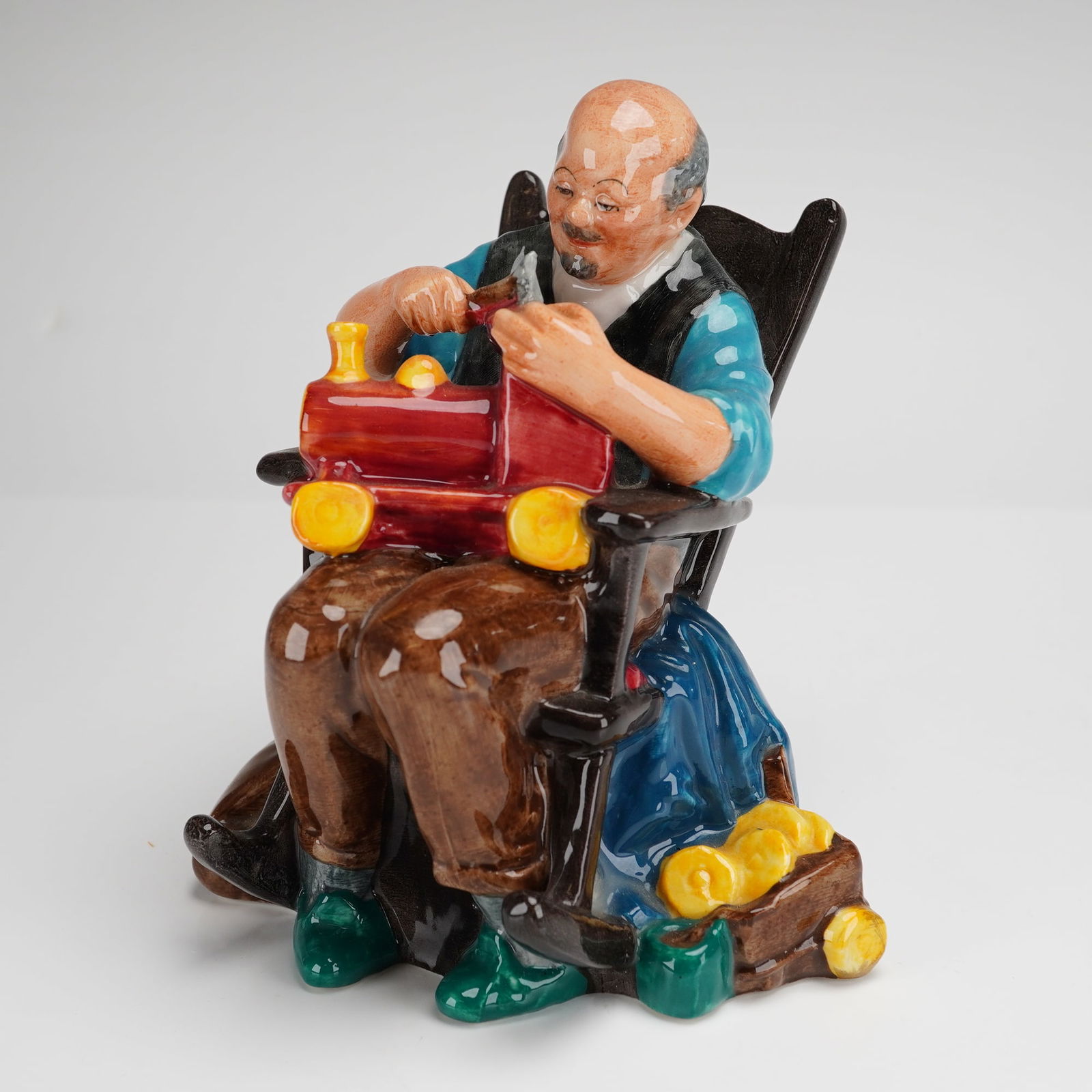 Royal Doulton Figurine The Toymaker HN2250: A Royal Doulton glazed ceramic figurine titled The Toymaker, model number HN2250. The figure depicts an elderly artisan seated in a dark-colored rocking chair, focused on a red toy locomotive. The art