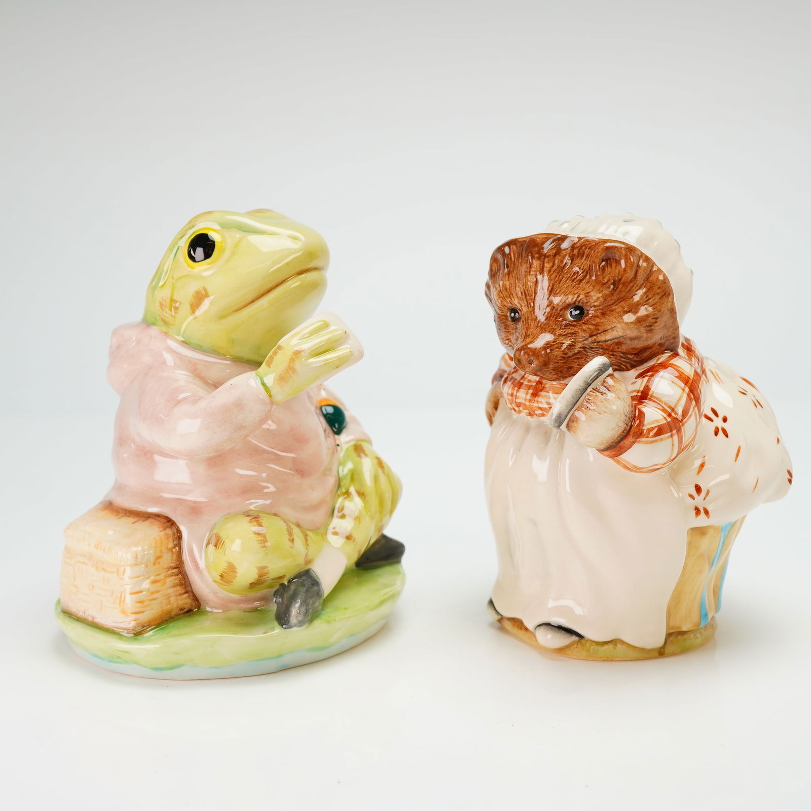 2 Royal Albert Beatrix Potter Figurines, Jeremy Fisher & Mrs. Tiggy Winkle: The glazed figures include the characters Jeremy Fisher and Mrs. Tiggy-Winkle. Jeremy Fisher is depicted seated in a pink coat, and Mrs. Tiggy-Winkle is shown in her signature apron and bonnet. Both p
