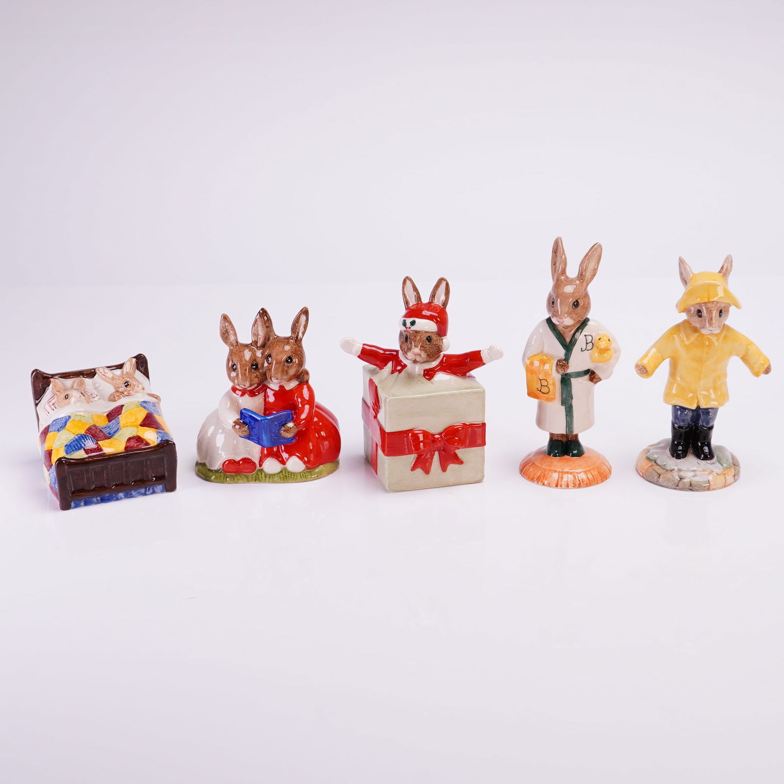 Collection of 5 Royal Doulton Bunnykins Figurines Including Rainy Day: A collection of five glazed ceramic figurines from the Royal Doulton Bunnykins line. The collection features anthropomorphic rabbits engaged in various activities. The figures include a pair of rabbit